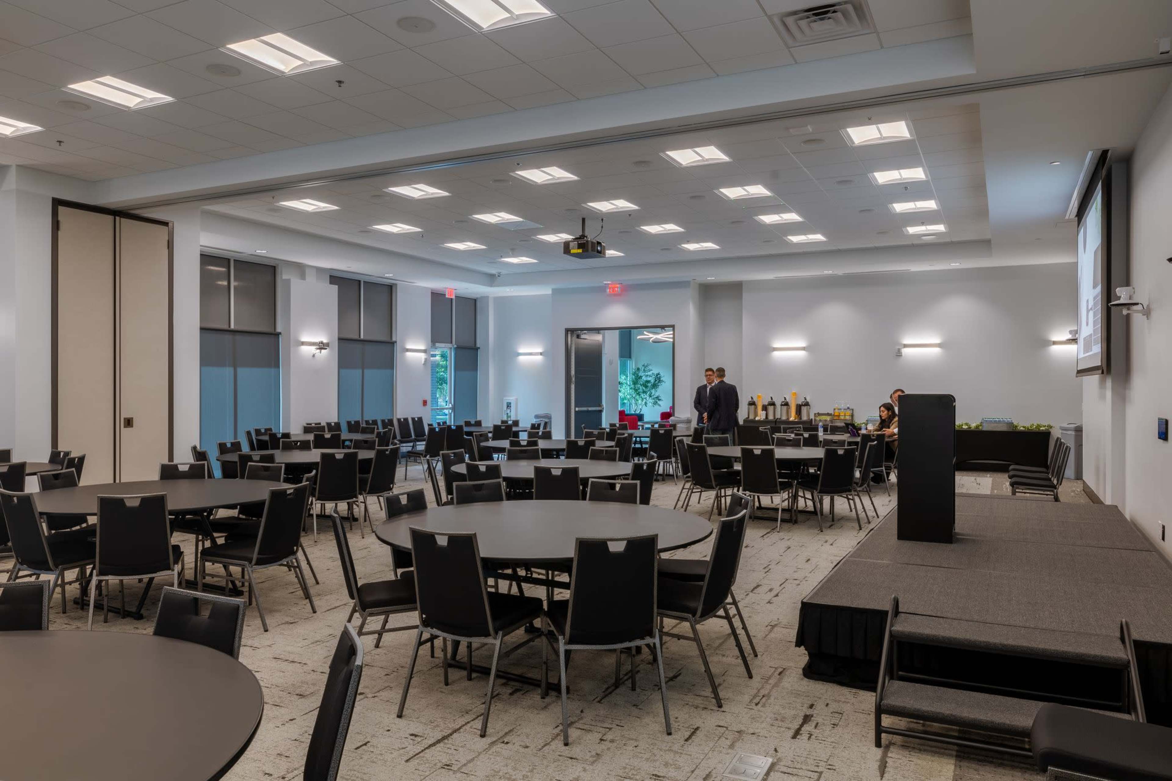 A spacious conference room features several round tables, a podium, and a refreshment area, with attendees conversing in the background.