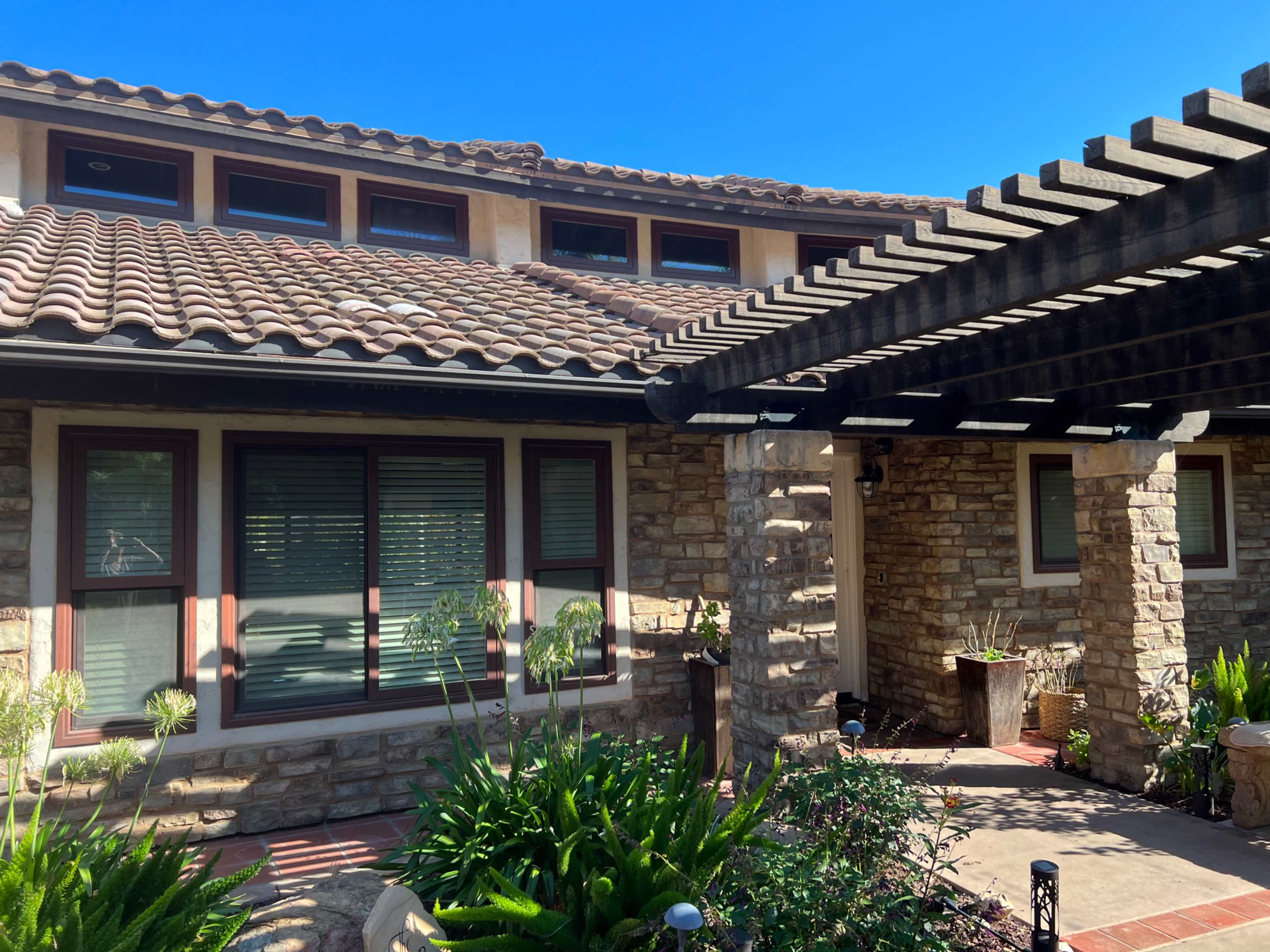 The image shows a house with a stone exterior, a tiled roof, and a pergola over a patio, surrounded by greenery.