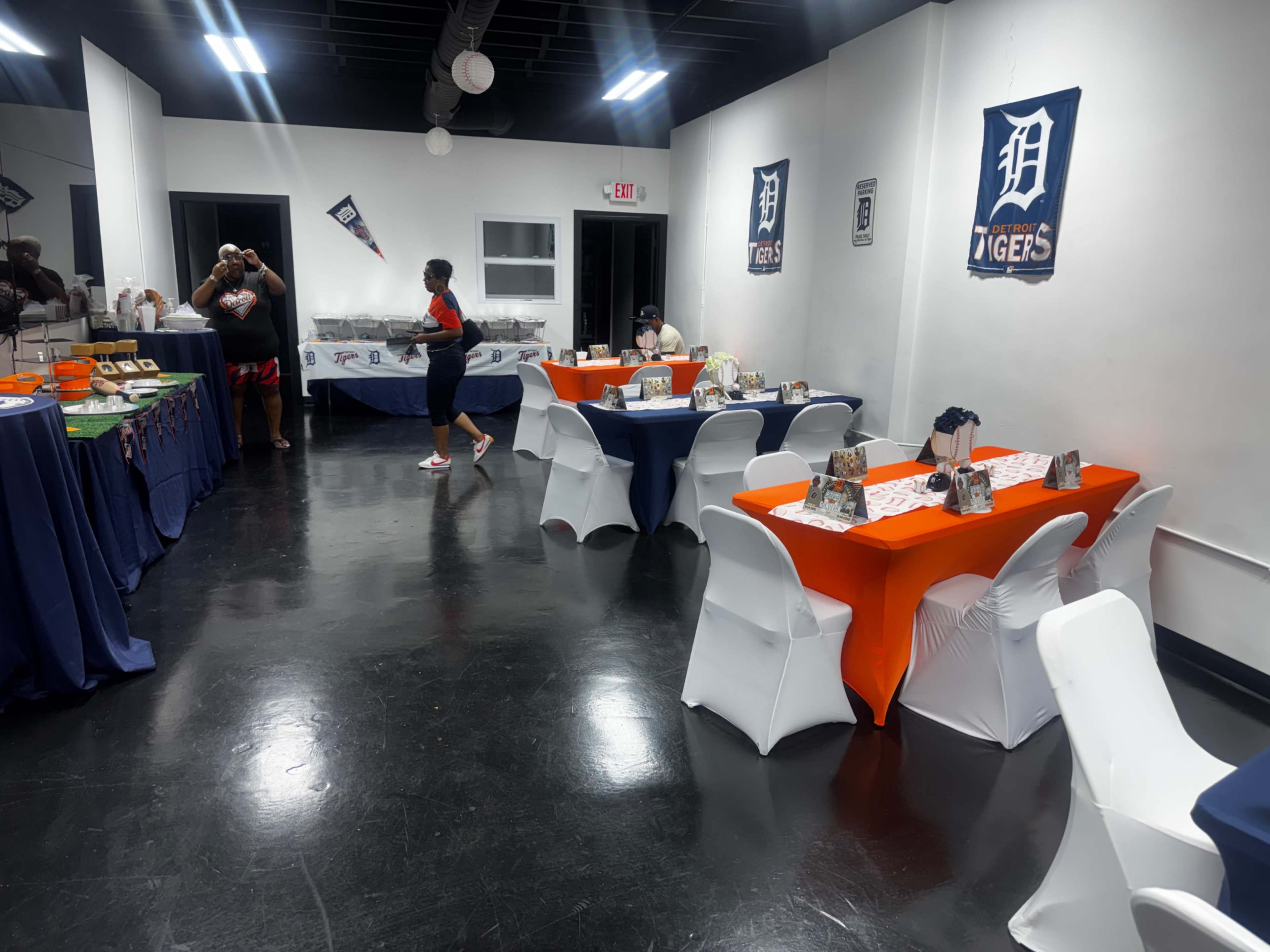 The image shows a spacious room set up for an event, featuring tables with orange and blue tablecloths, white chairs, and an array of food options displayed along the back wall.