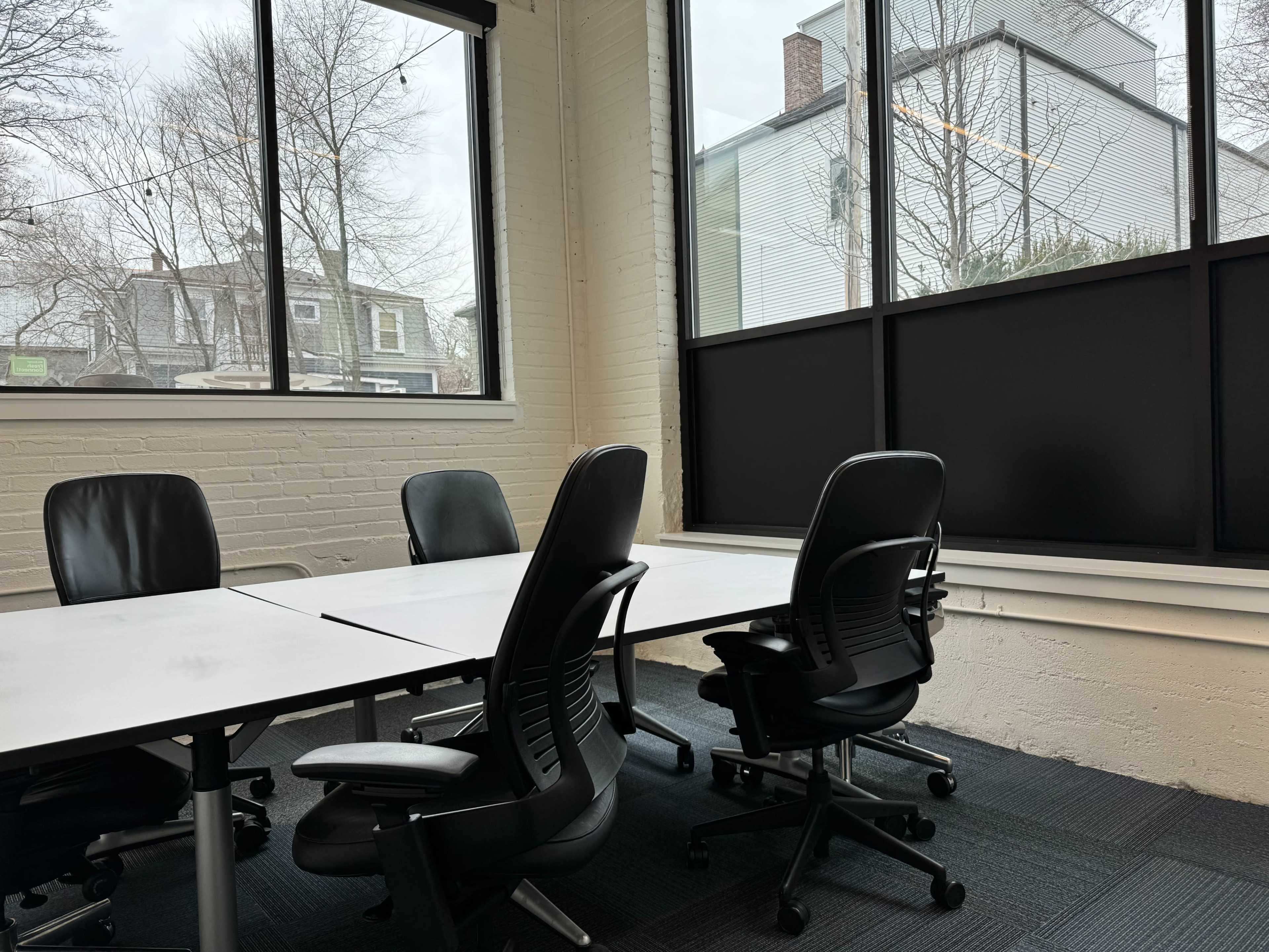 A conference room with four black chairs surrounding a white table, featuring large windows that overlook a gray, tree-lined street.