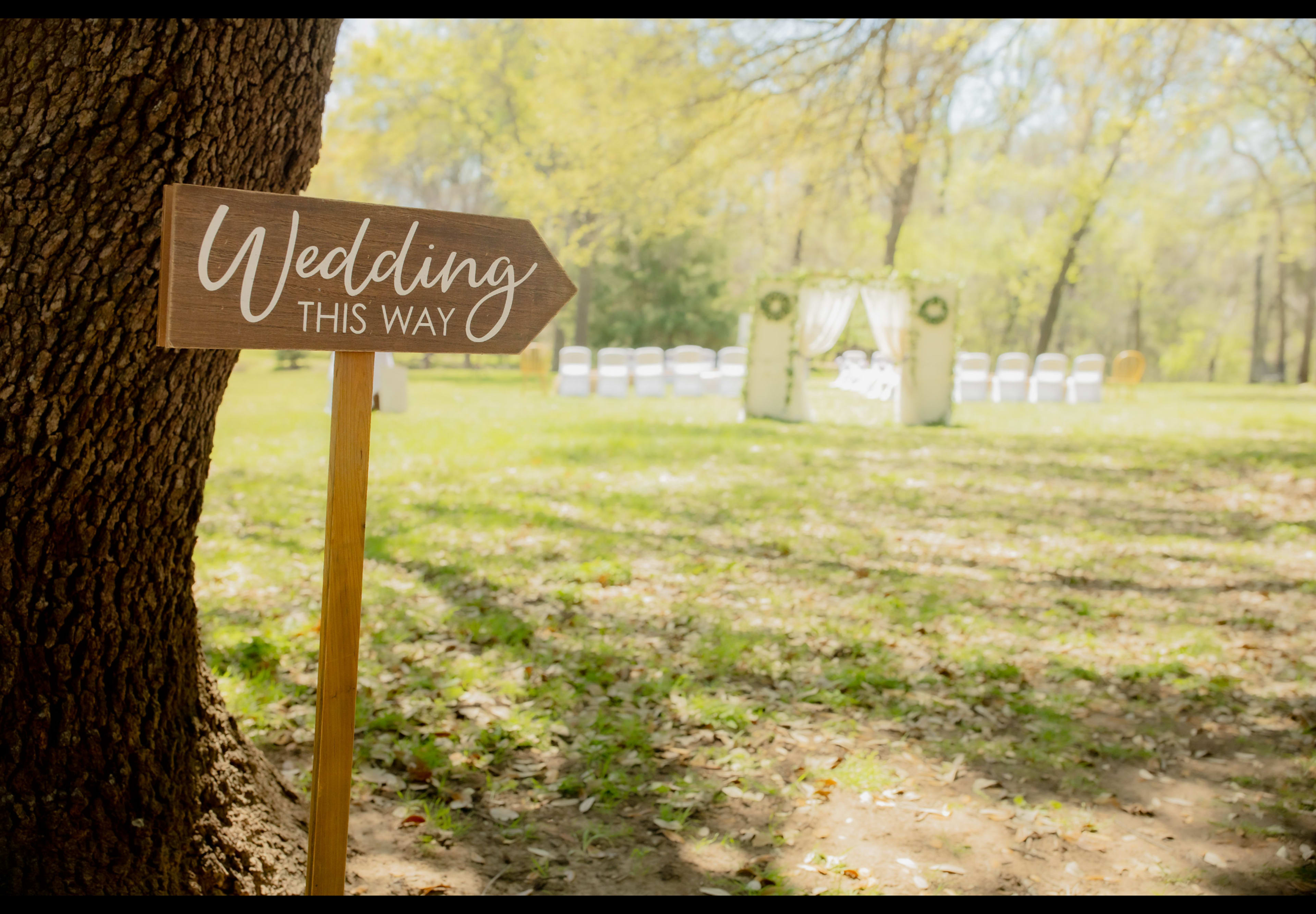 A wooden sign pointing towards a wedding setup in a grassy area features an arch and chairs arranged in a clearing.