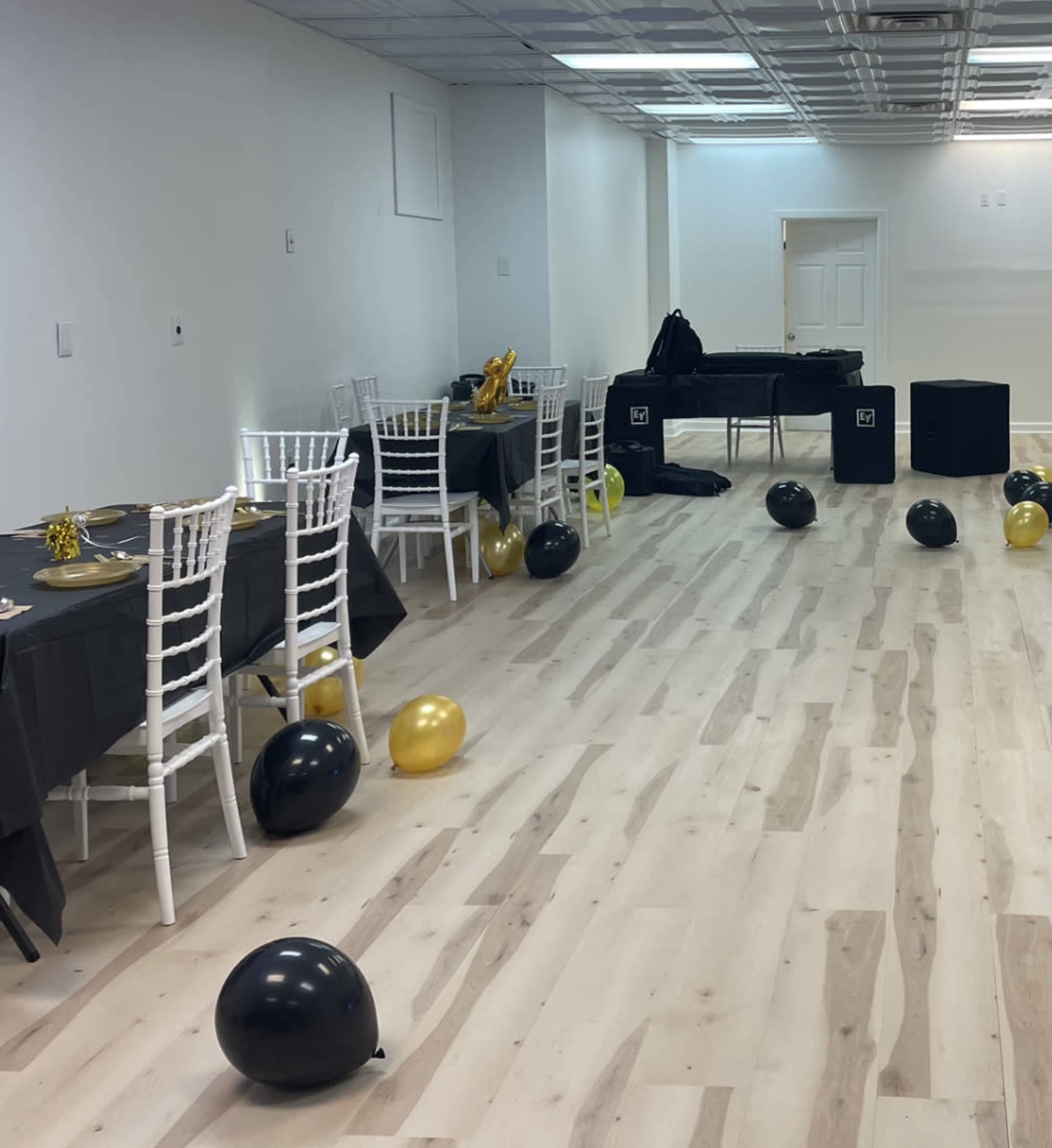 A brightly lit event space with tables set for a celebration, featuring black and gold décor and scattered balloons on the floor.