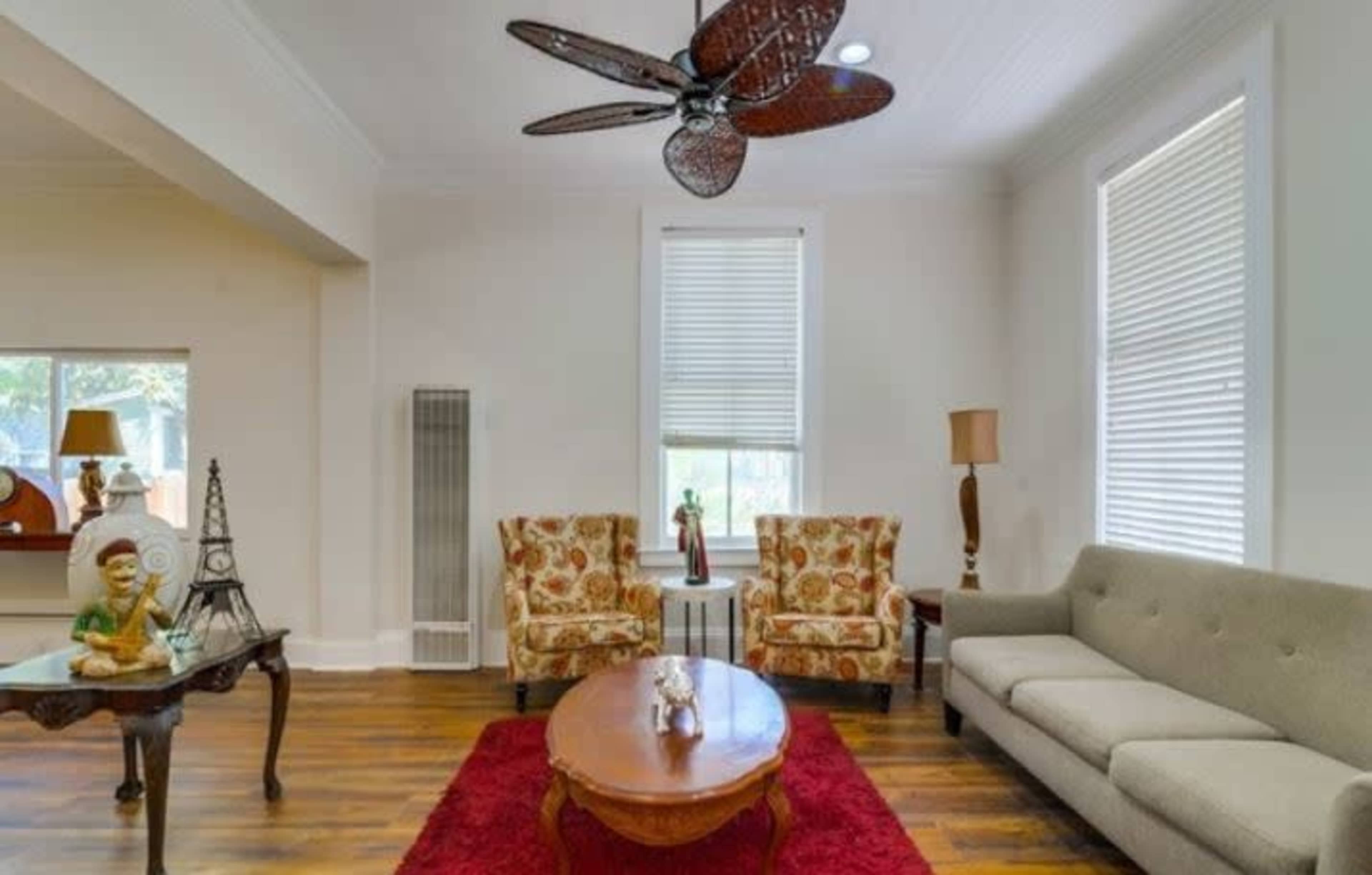 The living room features a gray sofa, two floral patterned chairs, a round wooden coffee table, and a ceiling fan above, against a backdrop of light-colored walls and hardwood flooring.