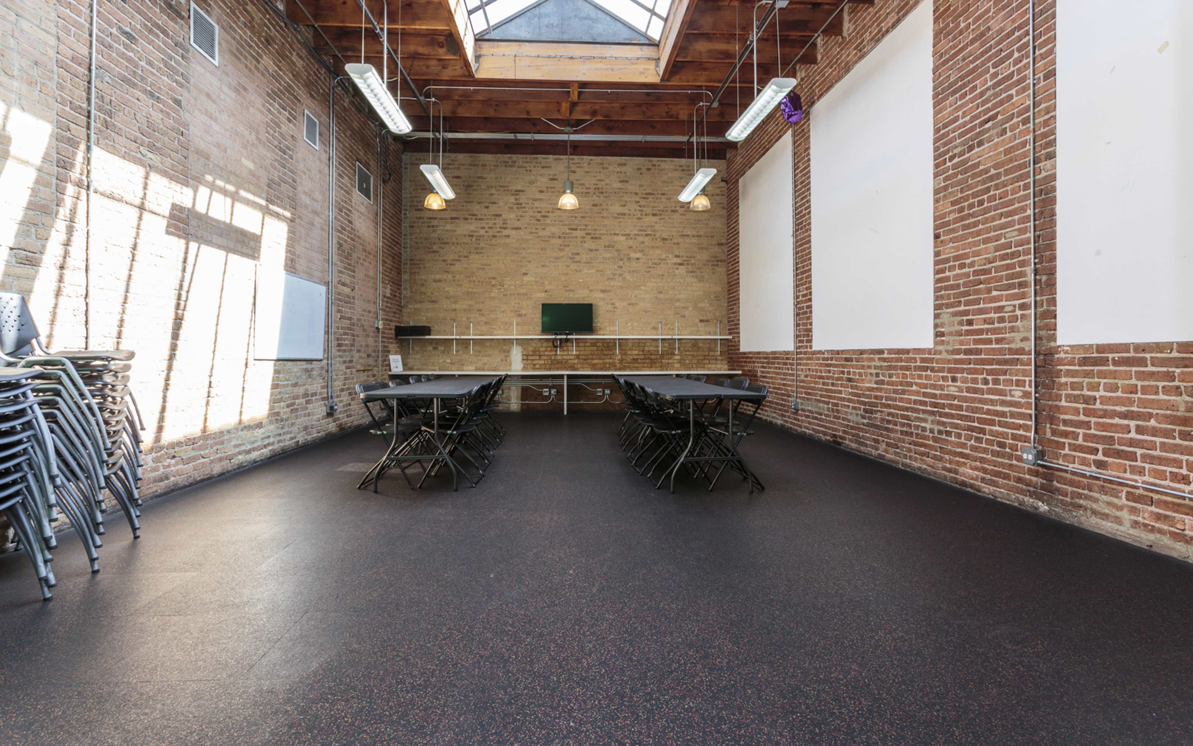 A spacious room with brick walls, a skylight, and several folding tables arranged in rows, along with stacked chairs.