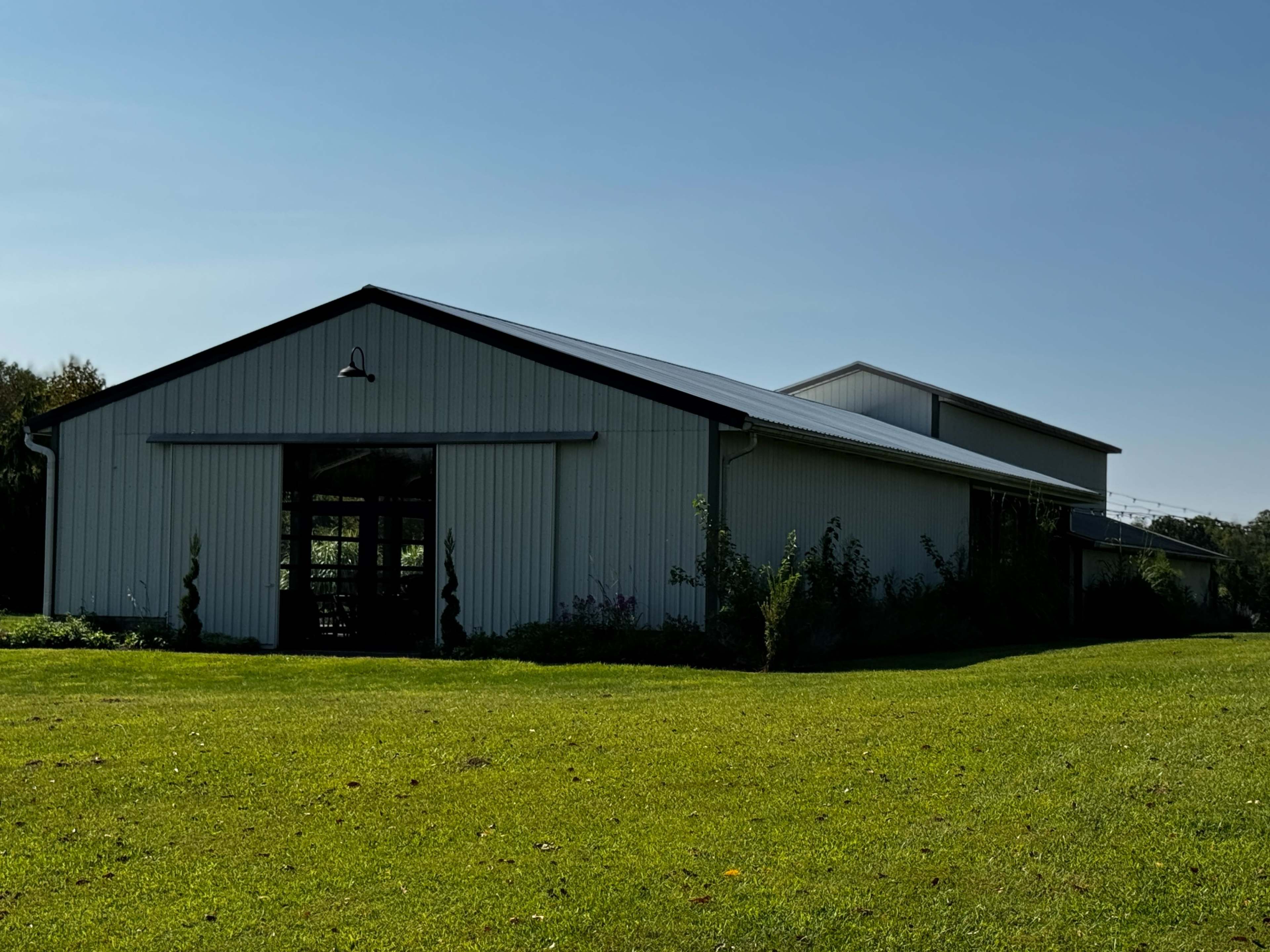 A large, metal-sided building with a sloped roof is situated on a grassy area under a clear blue sky.