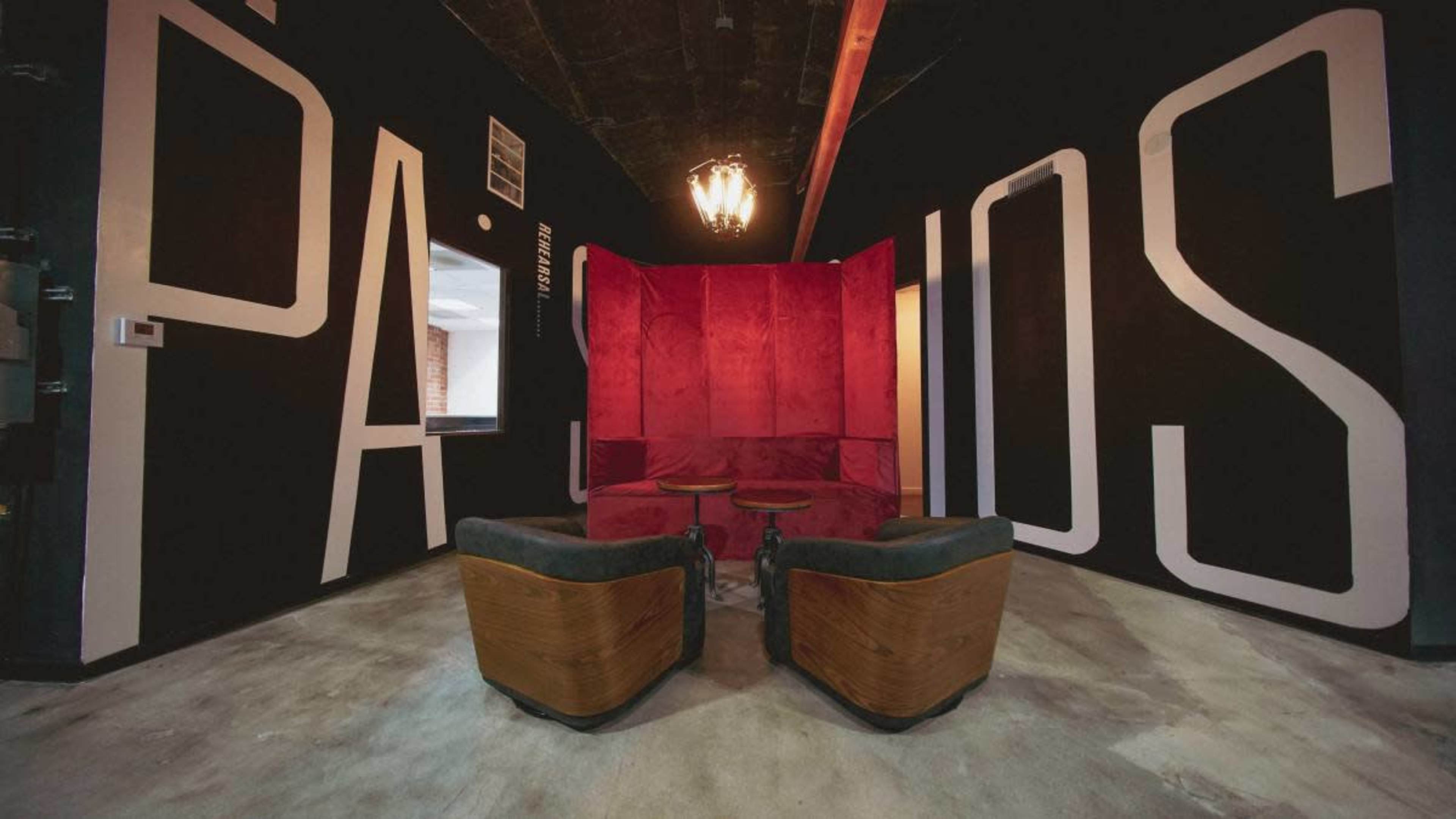 The image shows a room with a large black wall featuring the word "PASIONES" in white, and a seating area with two wooden chairs and a red velvet partition in the center.