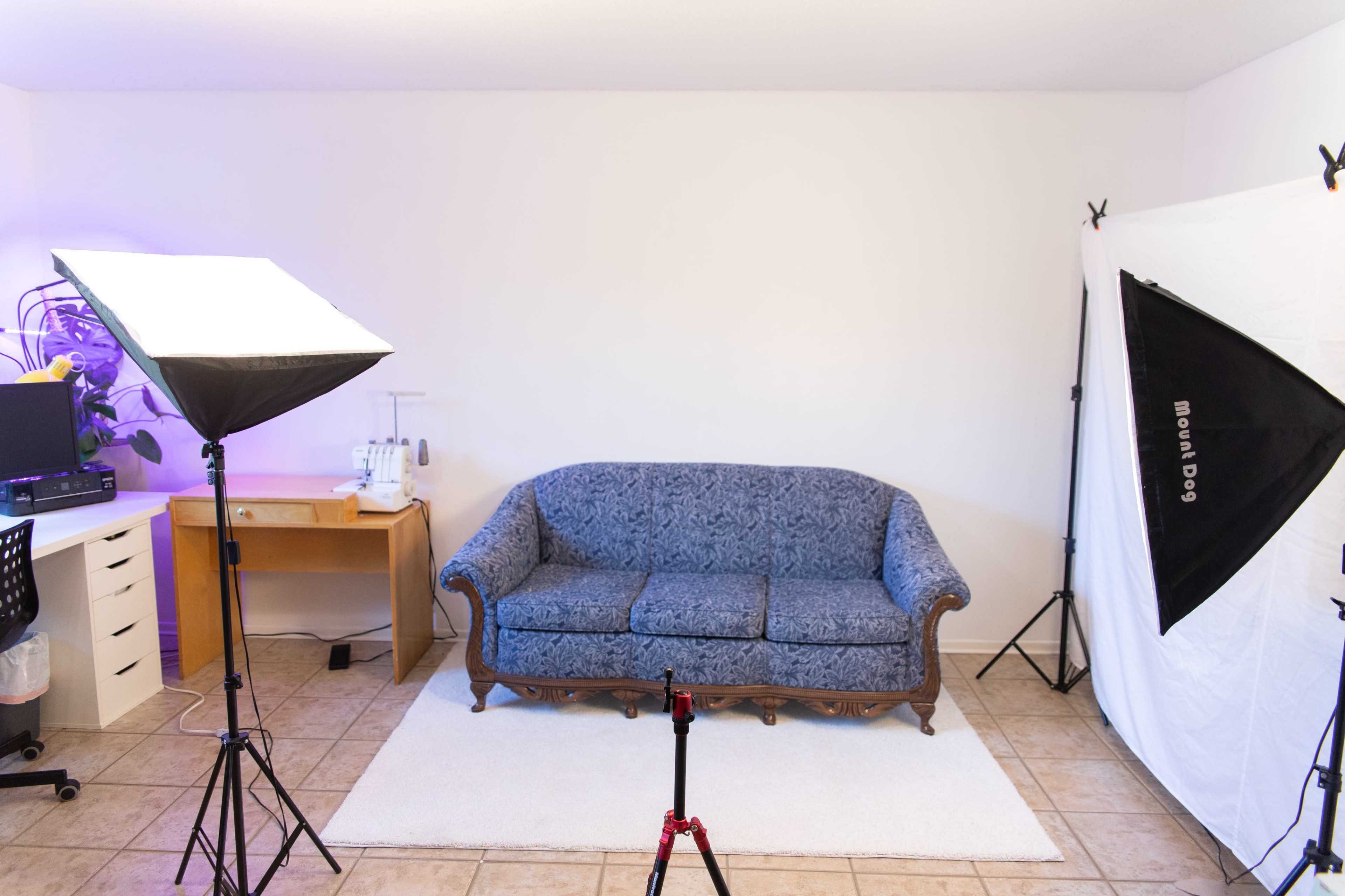 A well-lit photography studio featuring a blue sofa against a white wall, with two softbox lights and a wooden desk in the background.