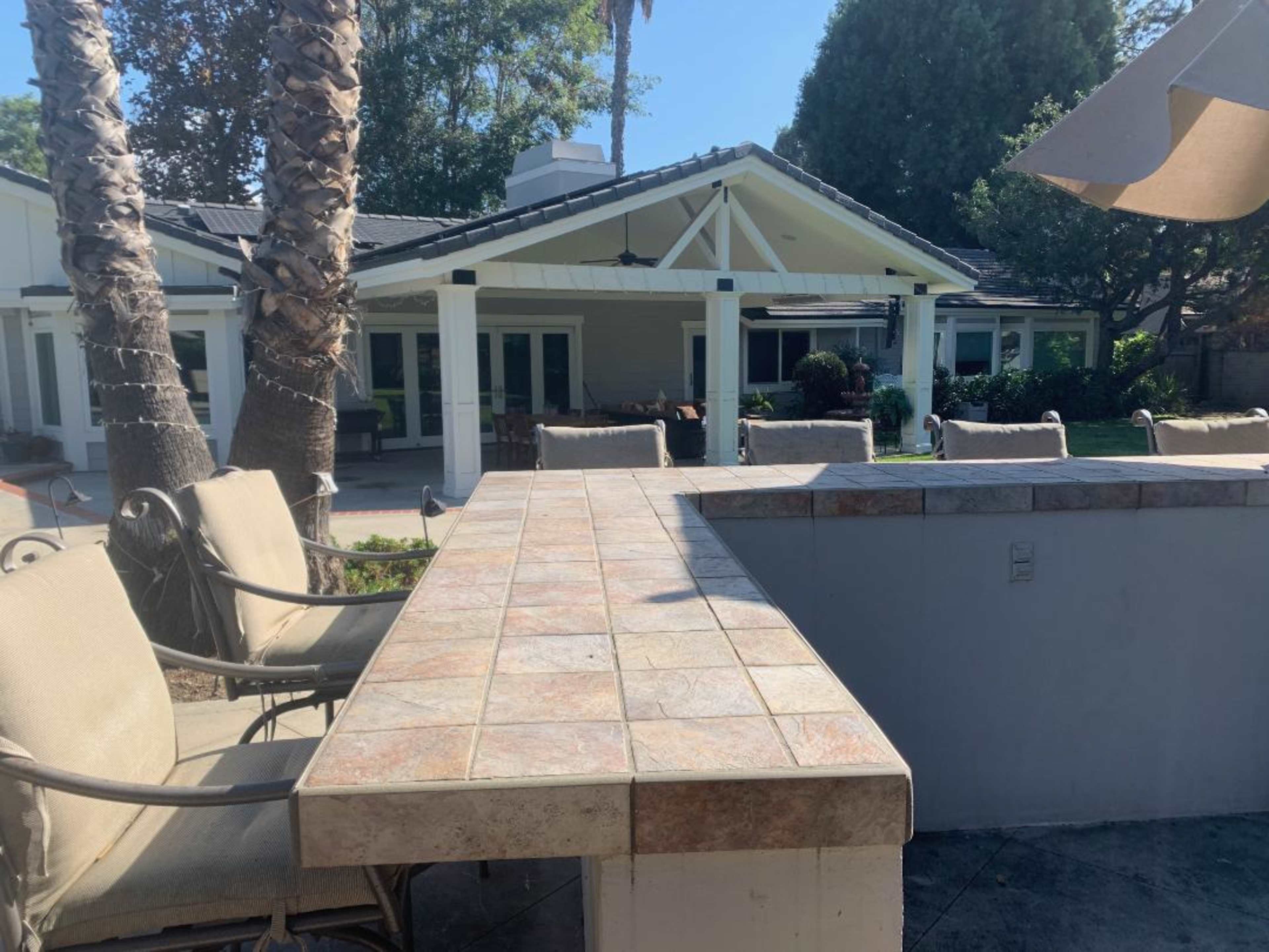 An outdoor patio area with a tiled bar, seating, and a view of a house surrounded by palm trees.