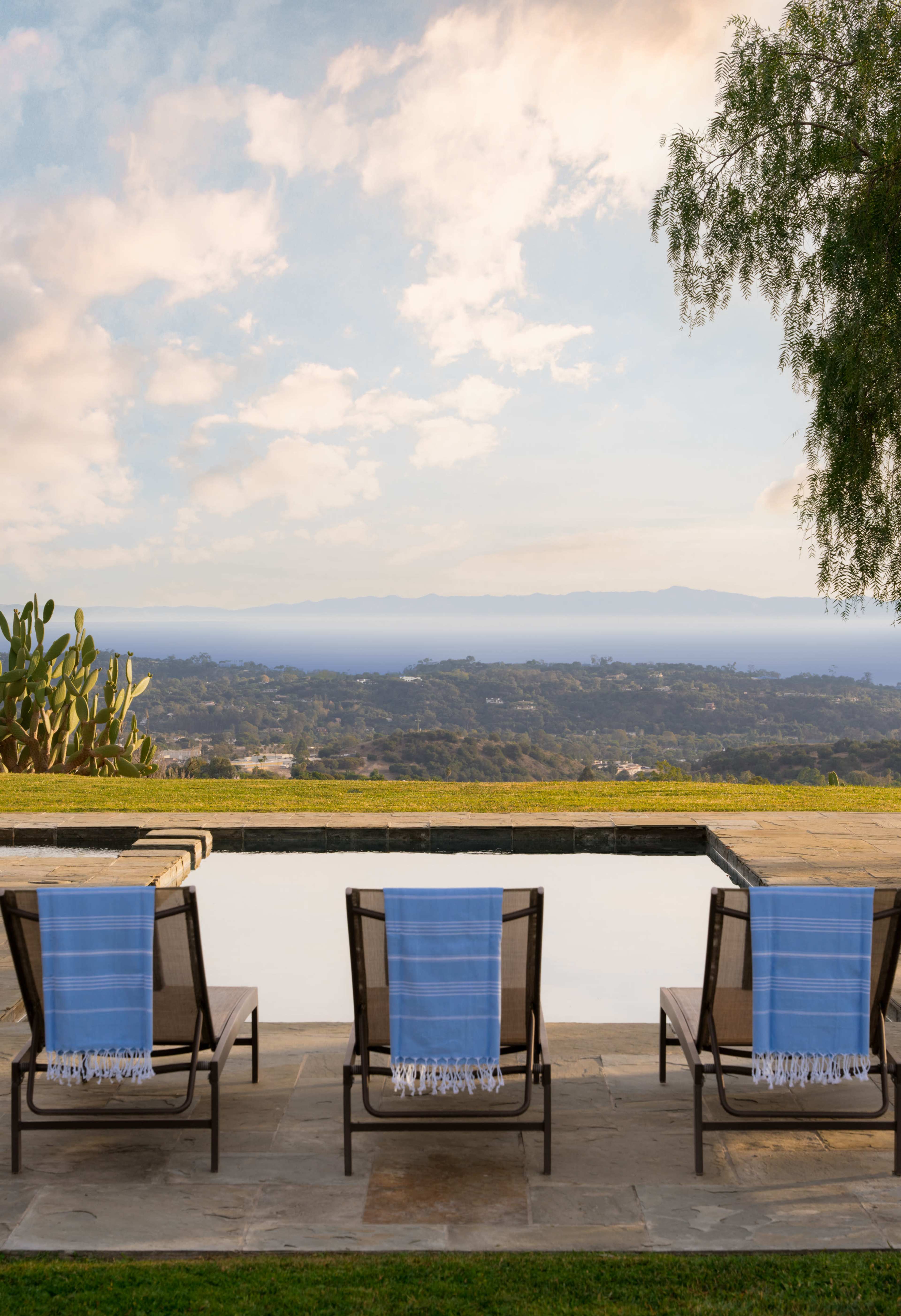 Lounge chairs with blue and white striped towels sit beside a pool that overlooks a green landscape and distant hills under a cloudy sky.
