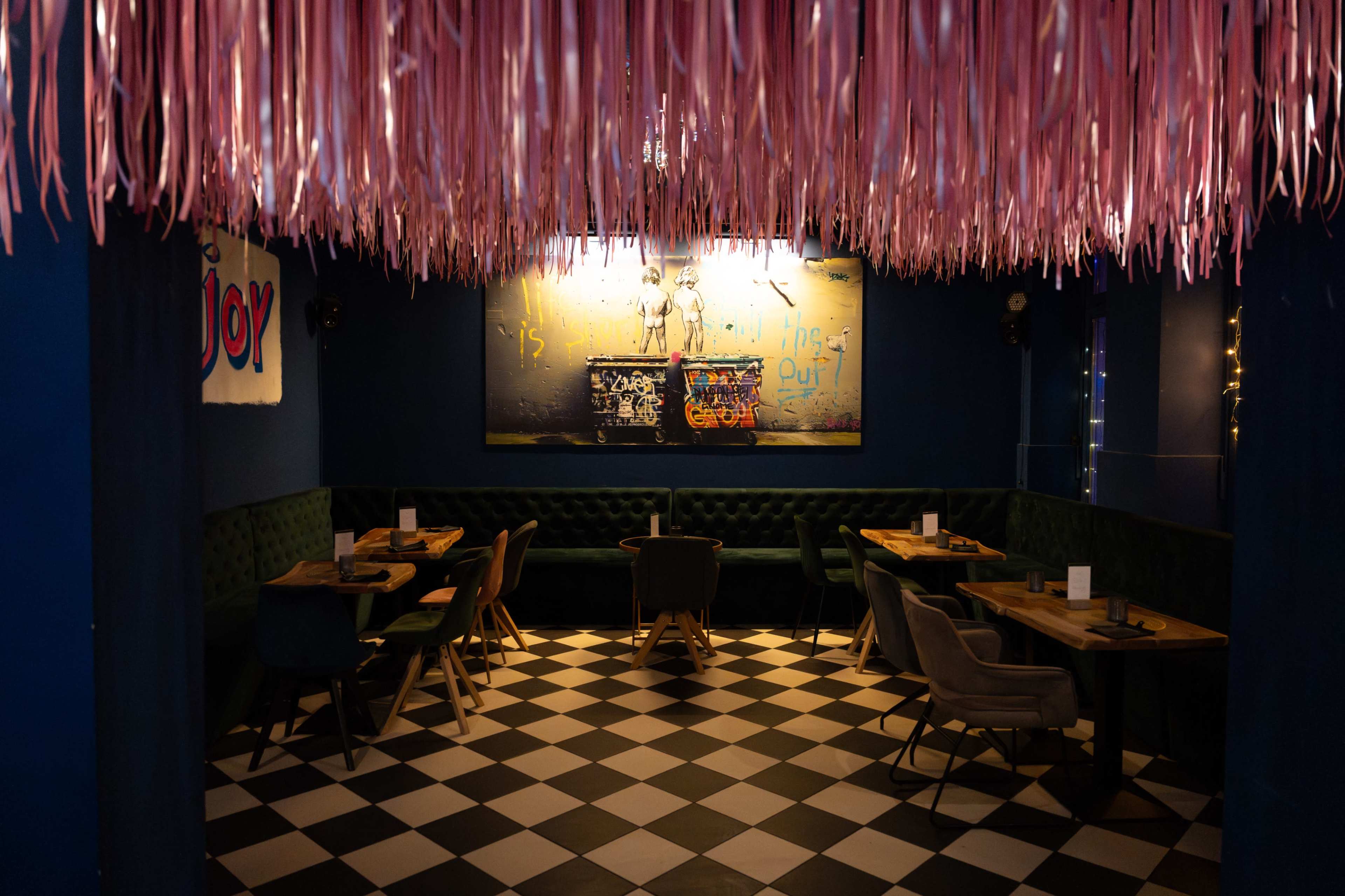 The image shows a restaurant interior with a checkered floor, green upholstered seating, wooden tables, and pink streamers hanging from the ceiling.