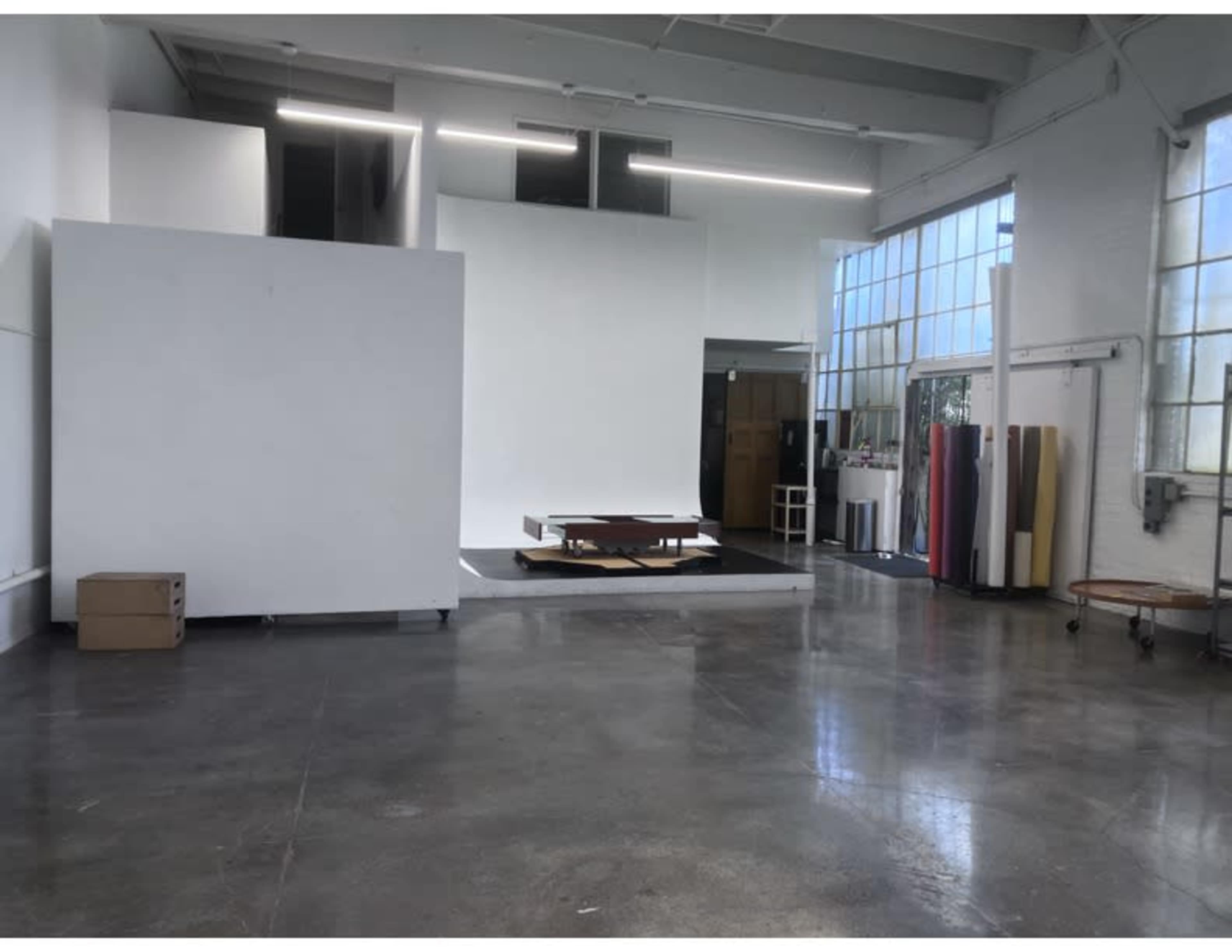 The image shows a spacious studio with a polished concrete floor, a large white backdrop, and a wooden platform in the center, surrounded by natural light from large windows.