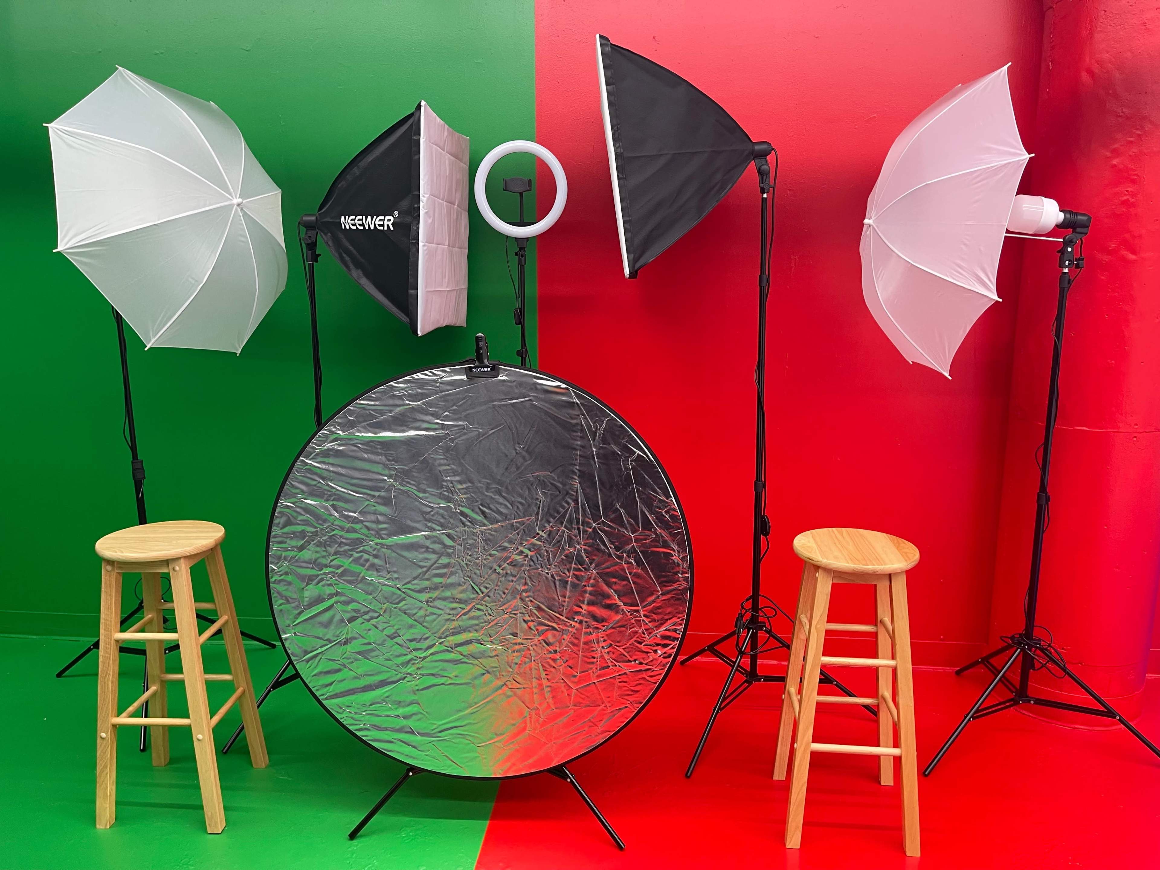 A photography setup with two wooden stools, three light stands with softboxes, a ring light, and a reflective silver disk against a red and green background.