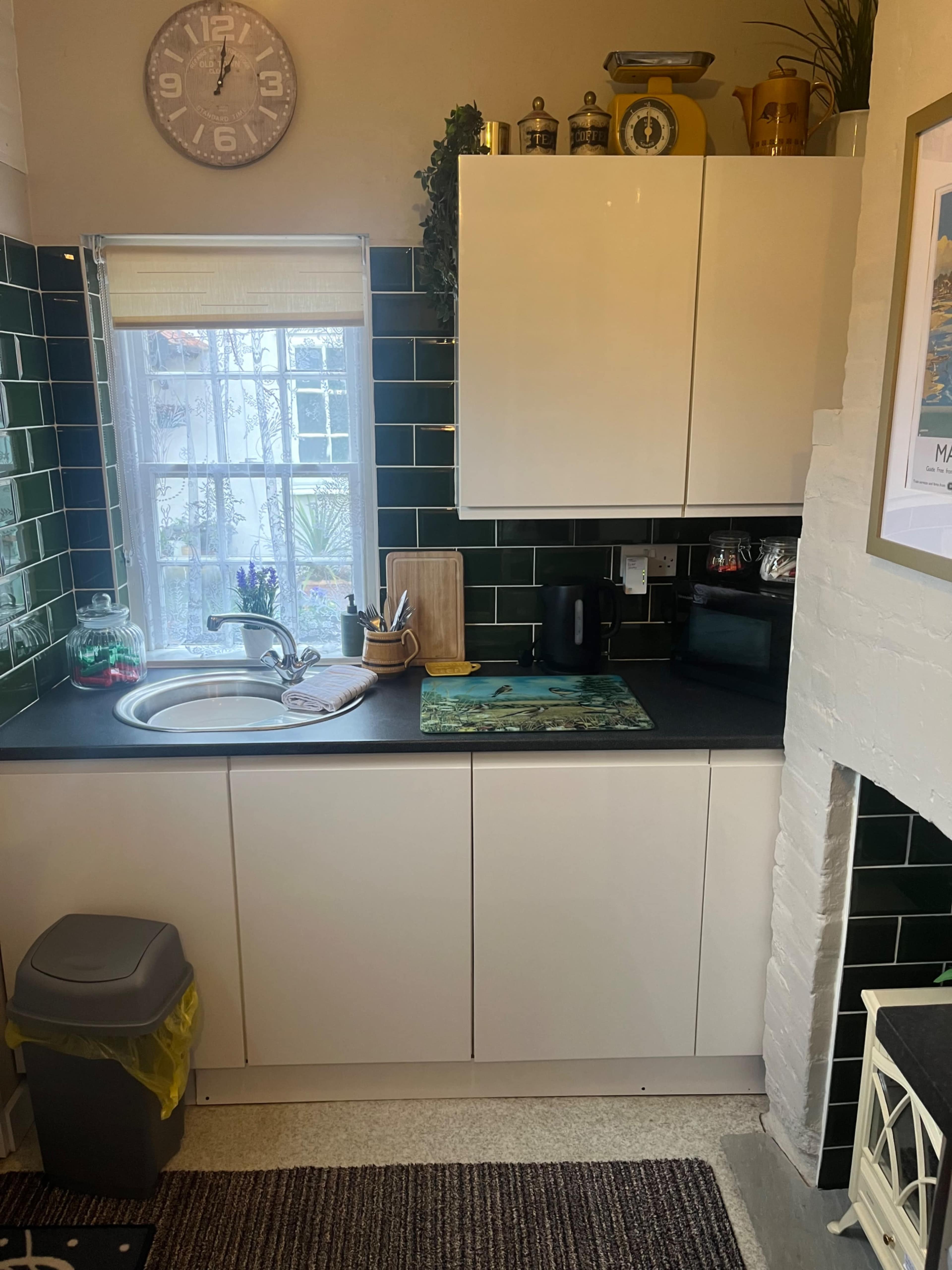 The image shows a small kitchen with green tiled walls, a sink, and white cabinets, featuring a kettle and cutting board on the counter.
