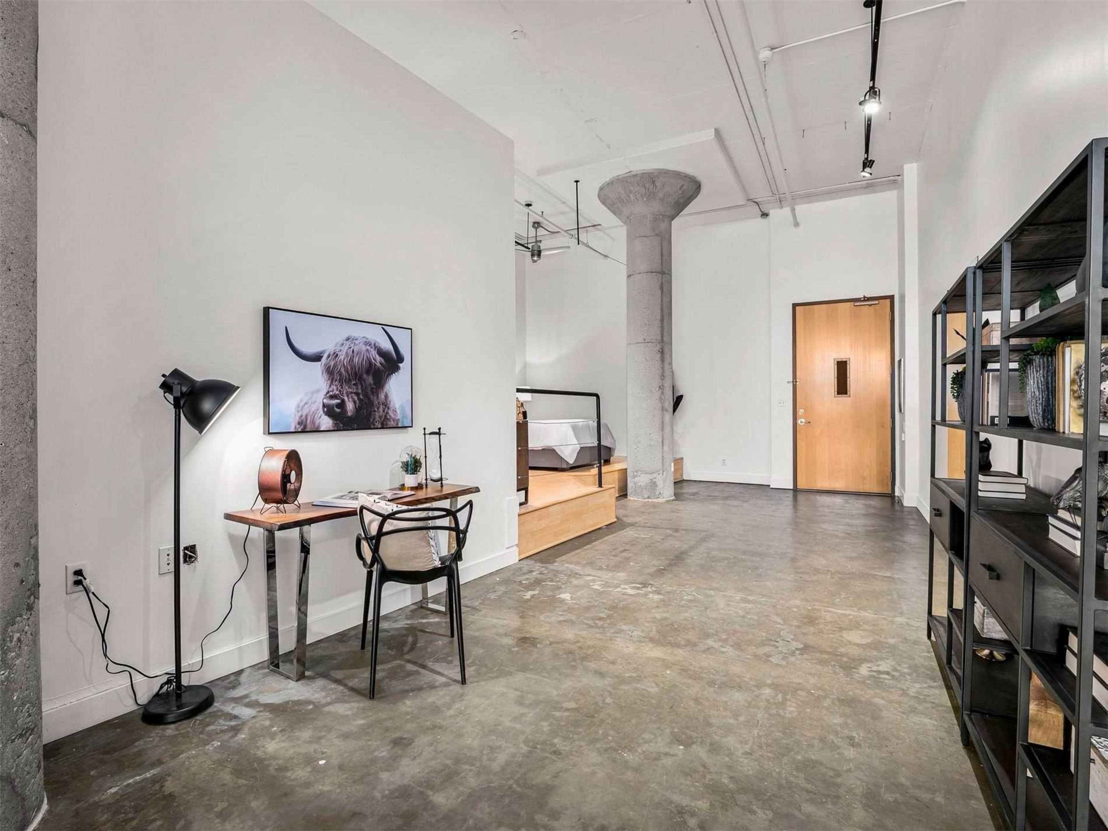 The image shows a modern, minimalistic studio apartment featuring a concrete floor, a desk with a chair, a bed on a raised platform, and a wall-mounted television.