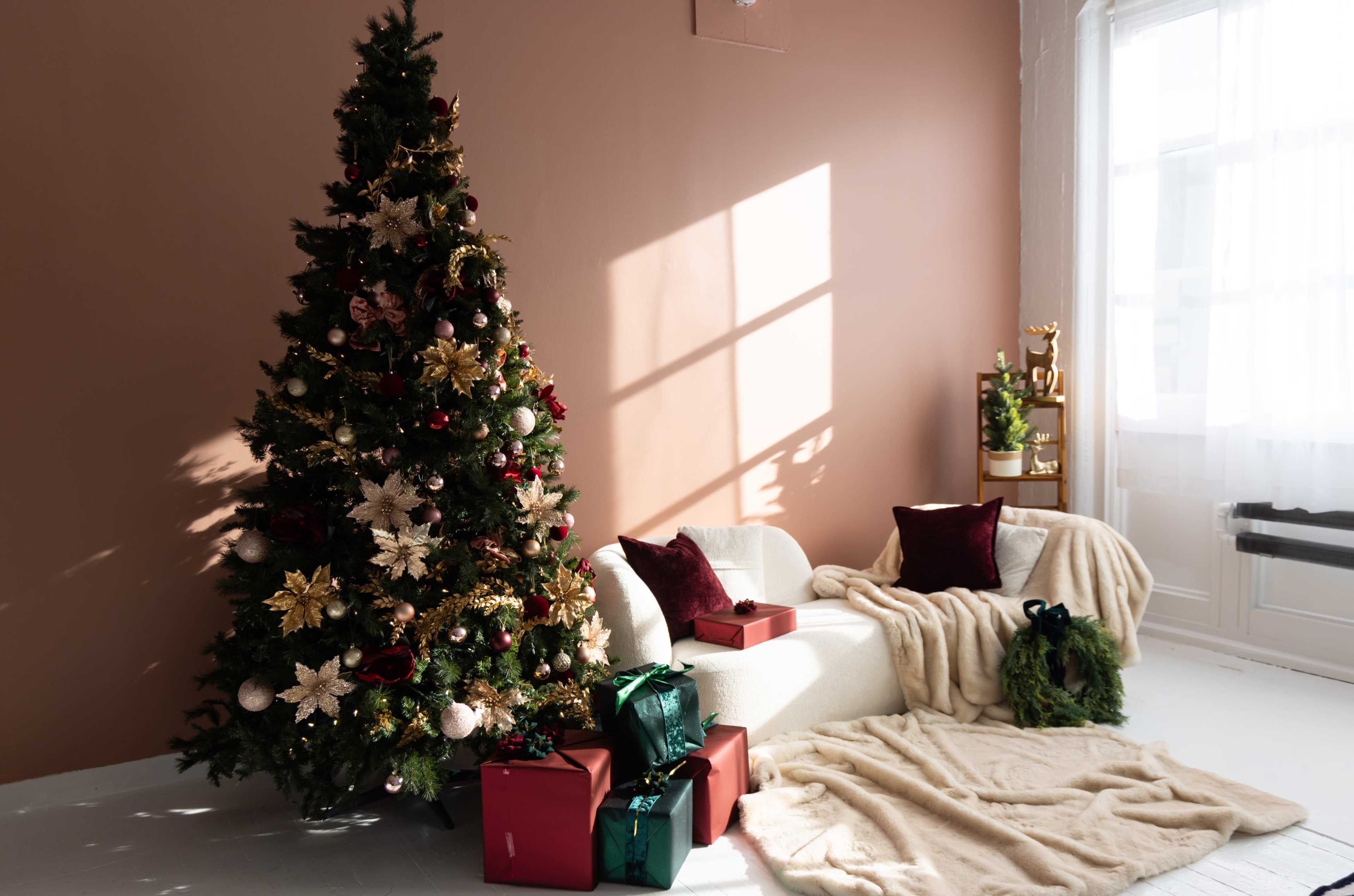 A decorated Christmas tree stands next to a white armchair draped with a blanket, surrounded by wrapped gifts in a softly lit room.