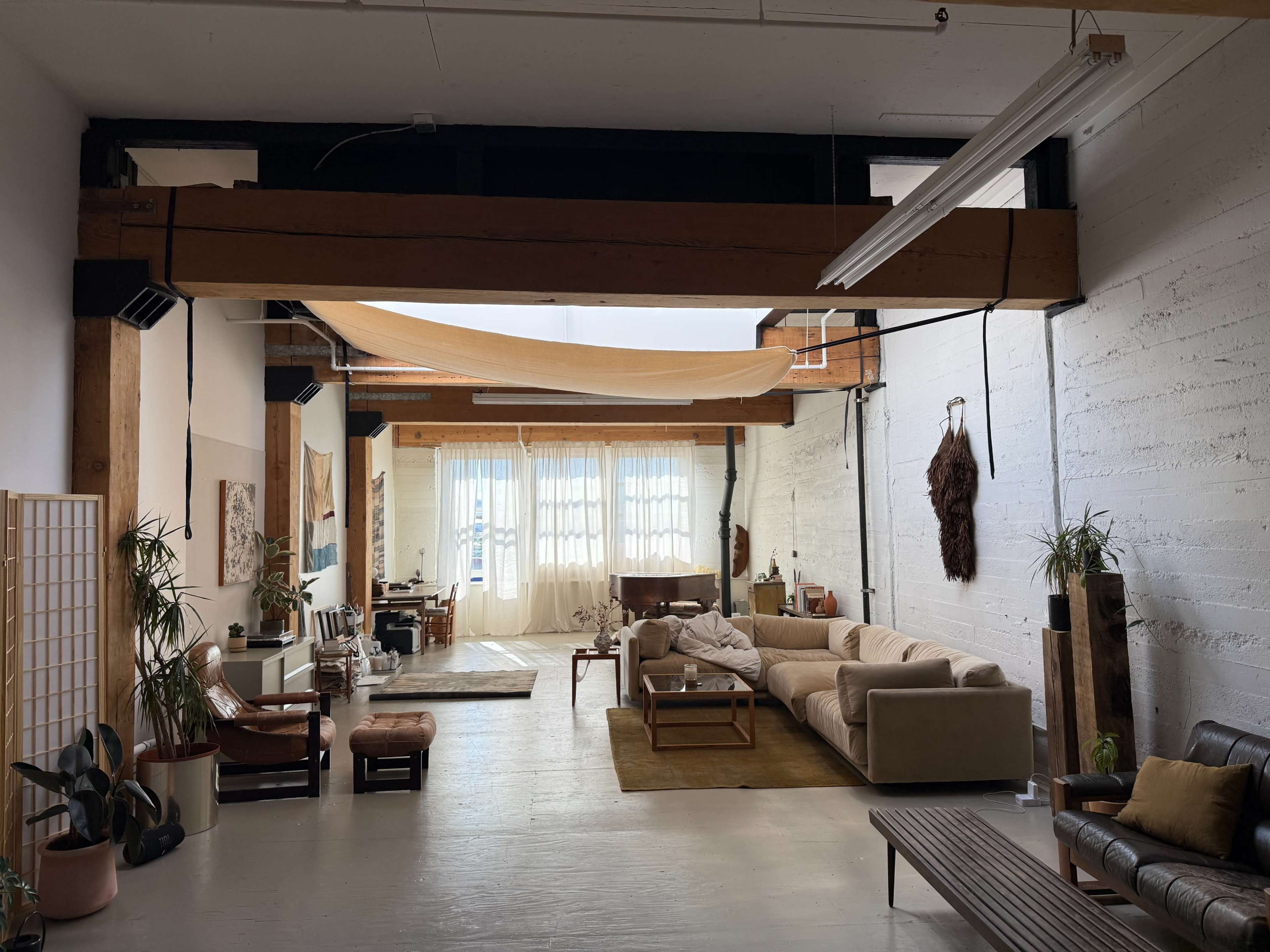 The image shows a spacious, minimalistic interior of a loft-style apartment with large windows, wooden beams, and various seating areas.