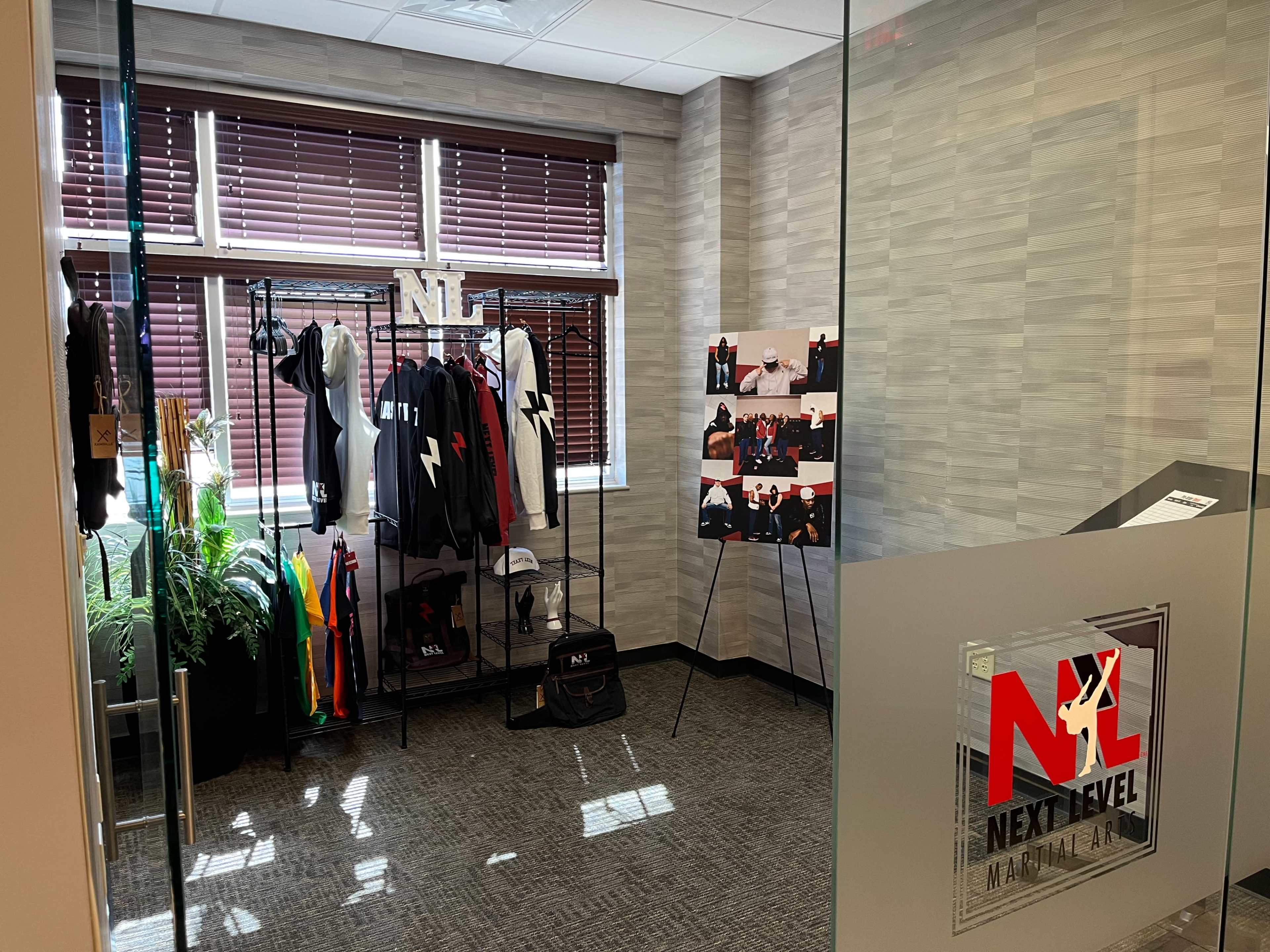 The image shows a showcase area with clothing racks displaying branded apparel and a poster board featuring product images, set in a well-lit room with glass walls.