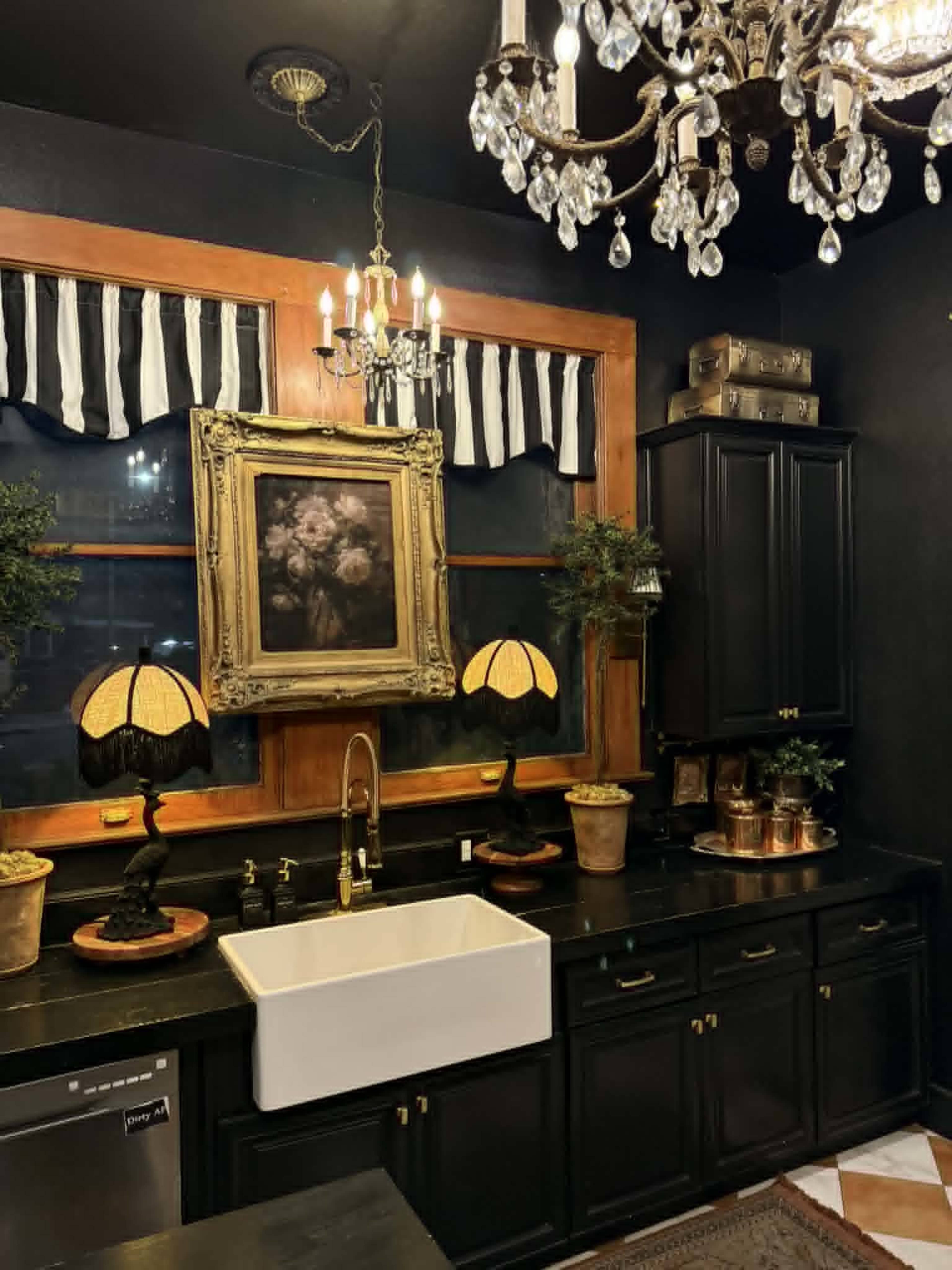 The image shows a dark-themed kitchen featuring a large farmhouse sink, decorative lamps on the counter, a framed painting on the wall, and a chandelier above.