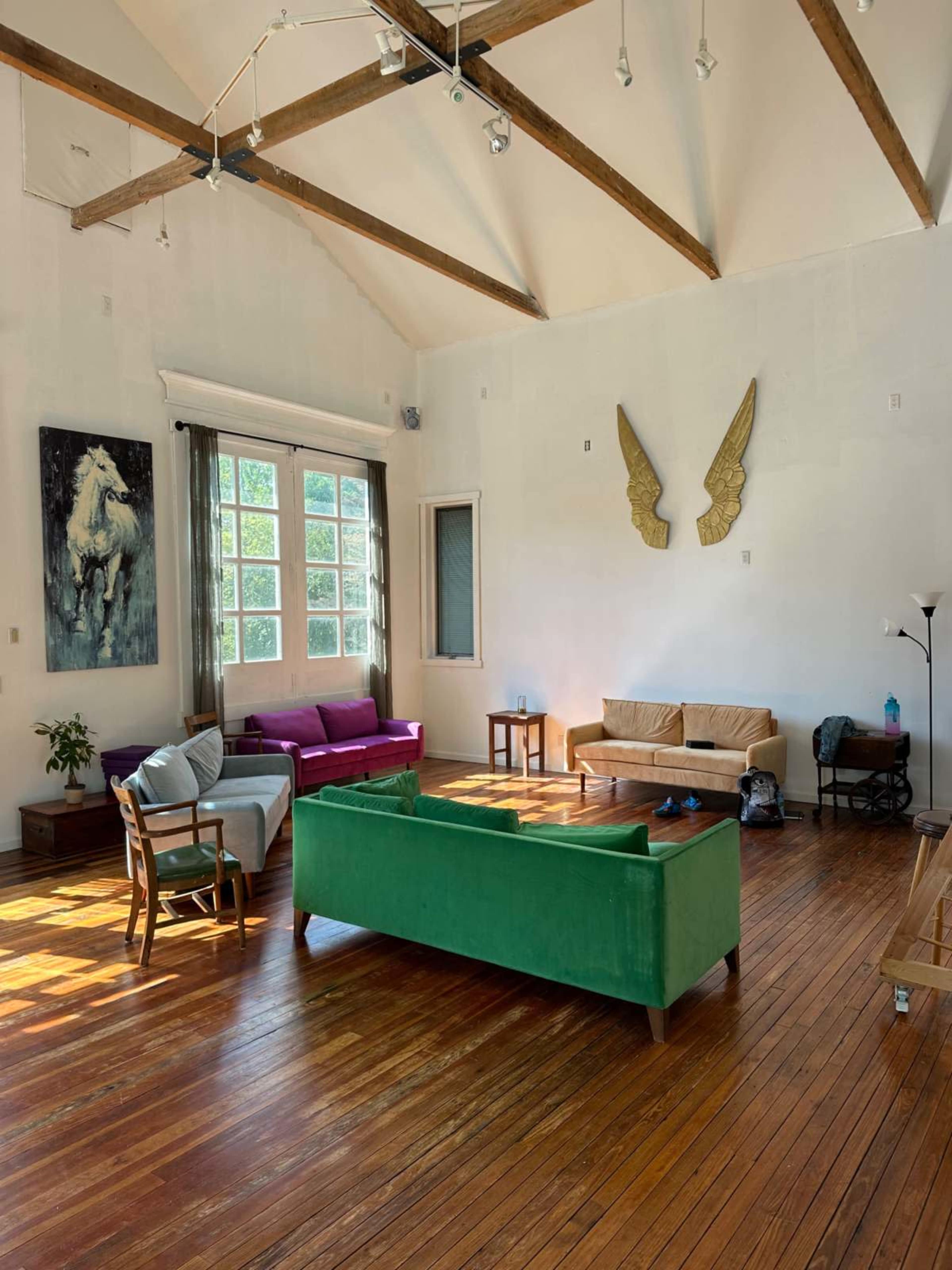 A spacious room with wooden floors features a green sofa, a pink sofa, and a wooden table, alongside large windows allowing ample natural light.