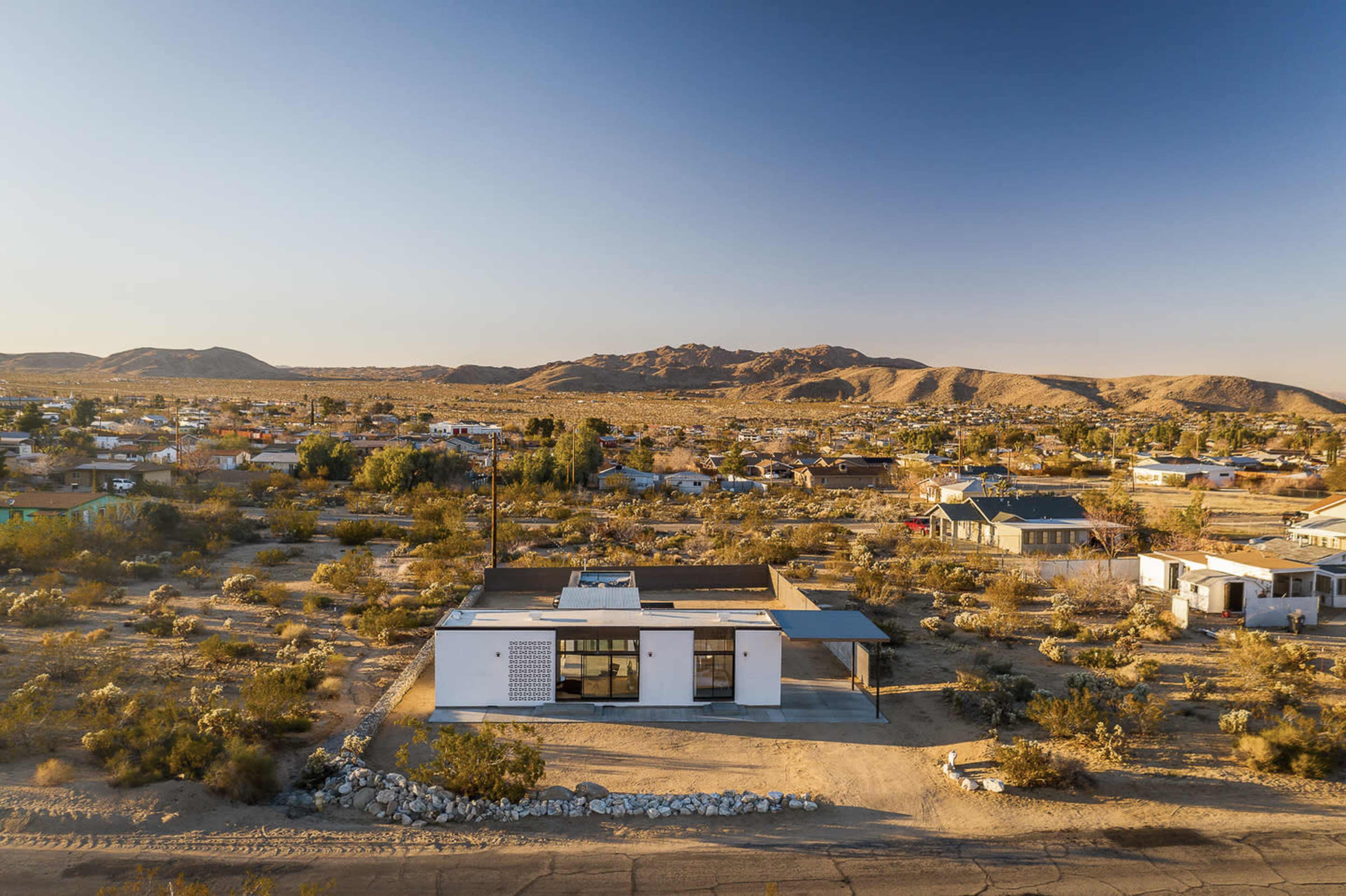 A modern white house stands among sparse vegetation and rocky terrain, with mountains in the distance and a small town visible nearby.