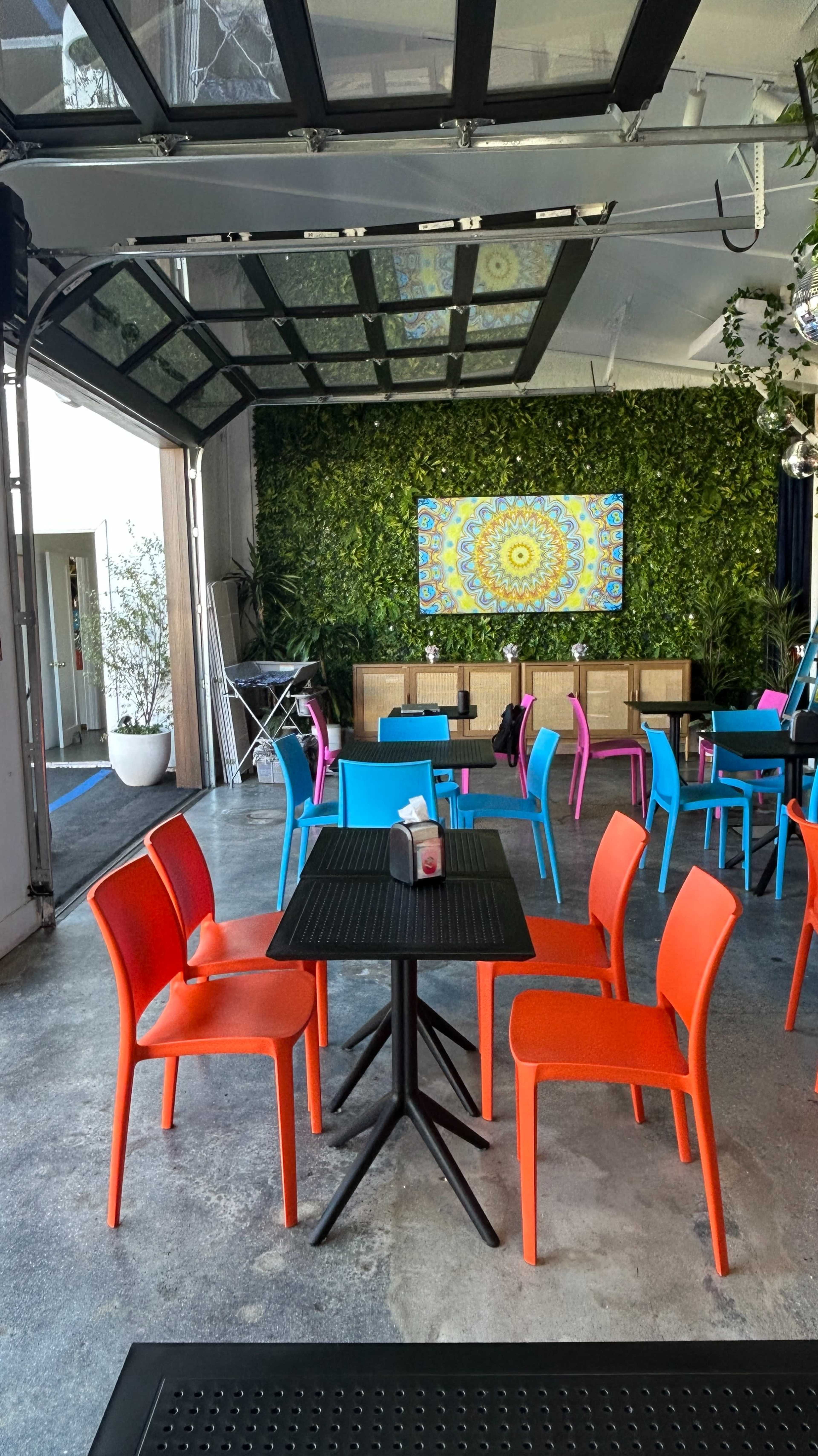 The image shows a modern café interior with vibrant chairs in pink, blue, and orange surrounding black tables, set against a green foliage wall and a colorful artwork.