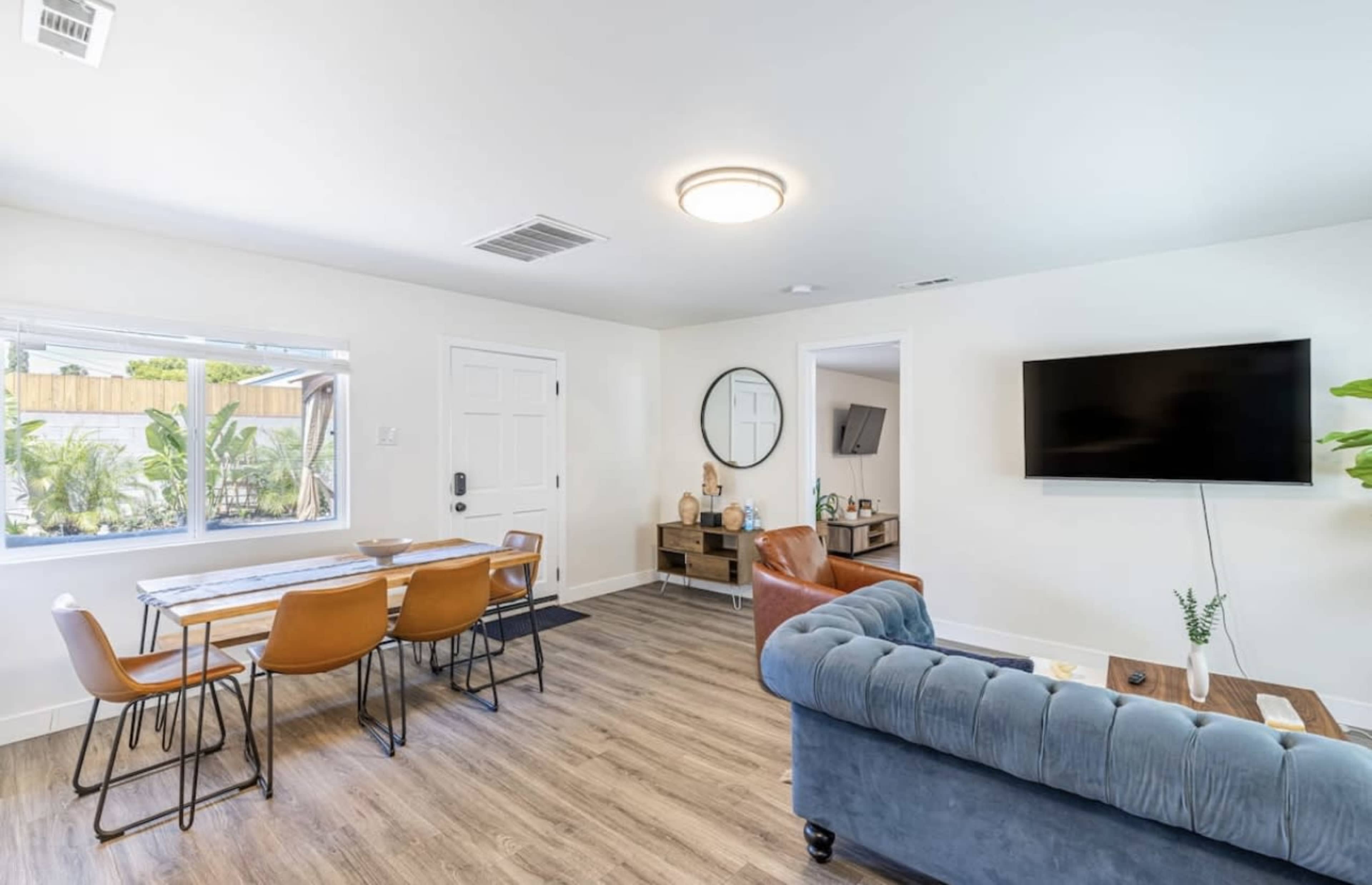 A modern living space featuring a dining table, a couch, a mounted television, and large windows letting in natural light.