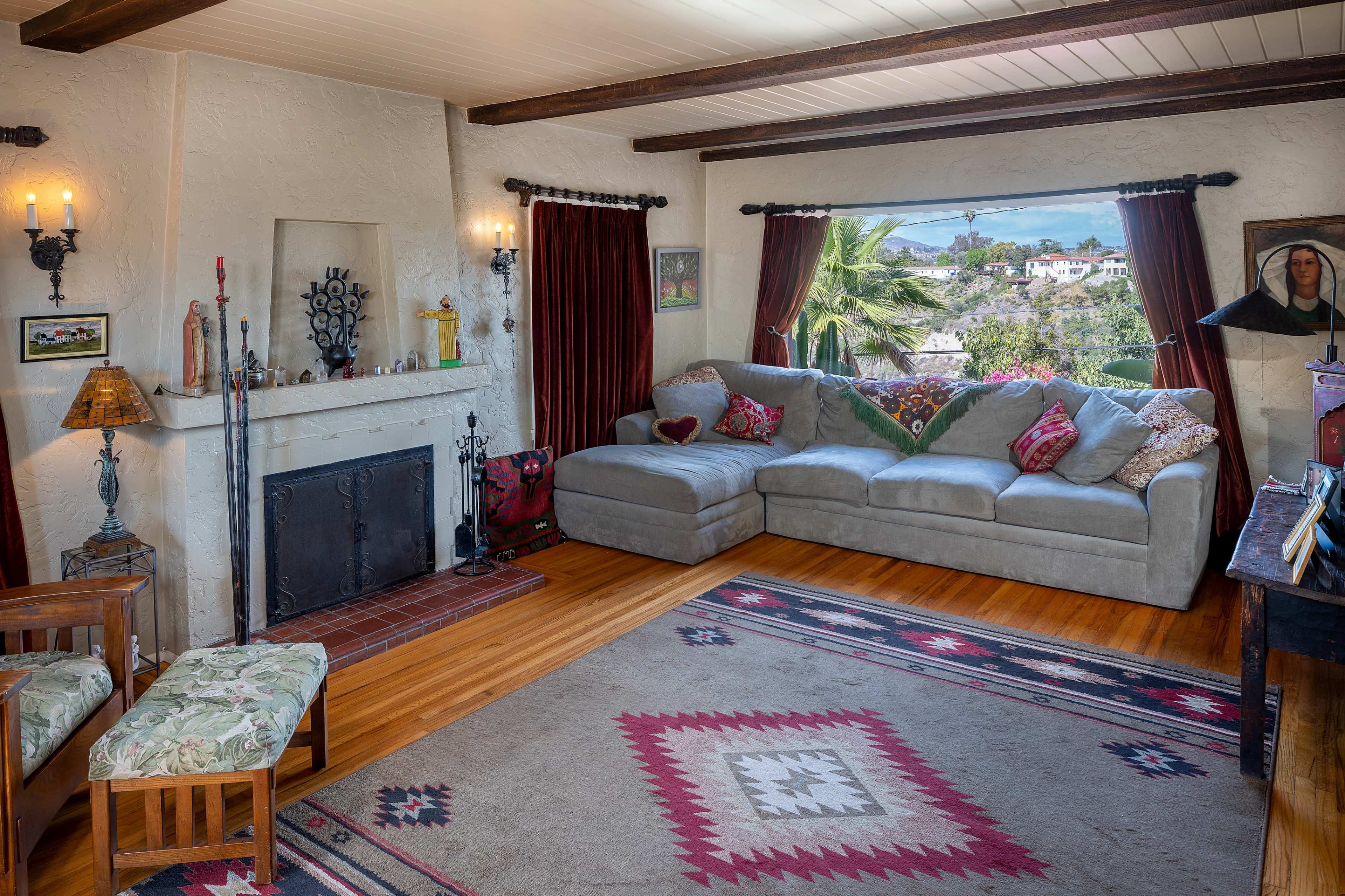 The room features a gray sectional sofa, a wooden coffee table, a fireplace, and large windows with drapes, all set against a backdrop of rustic decor and a patterned rug.