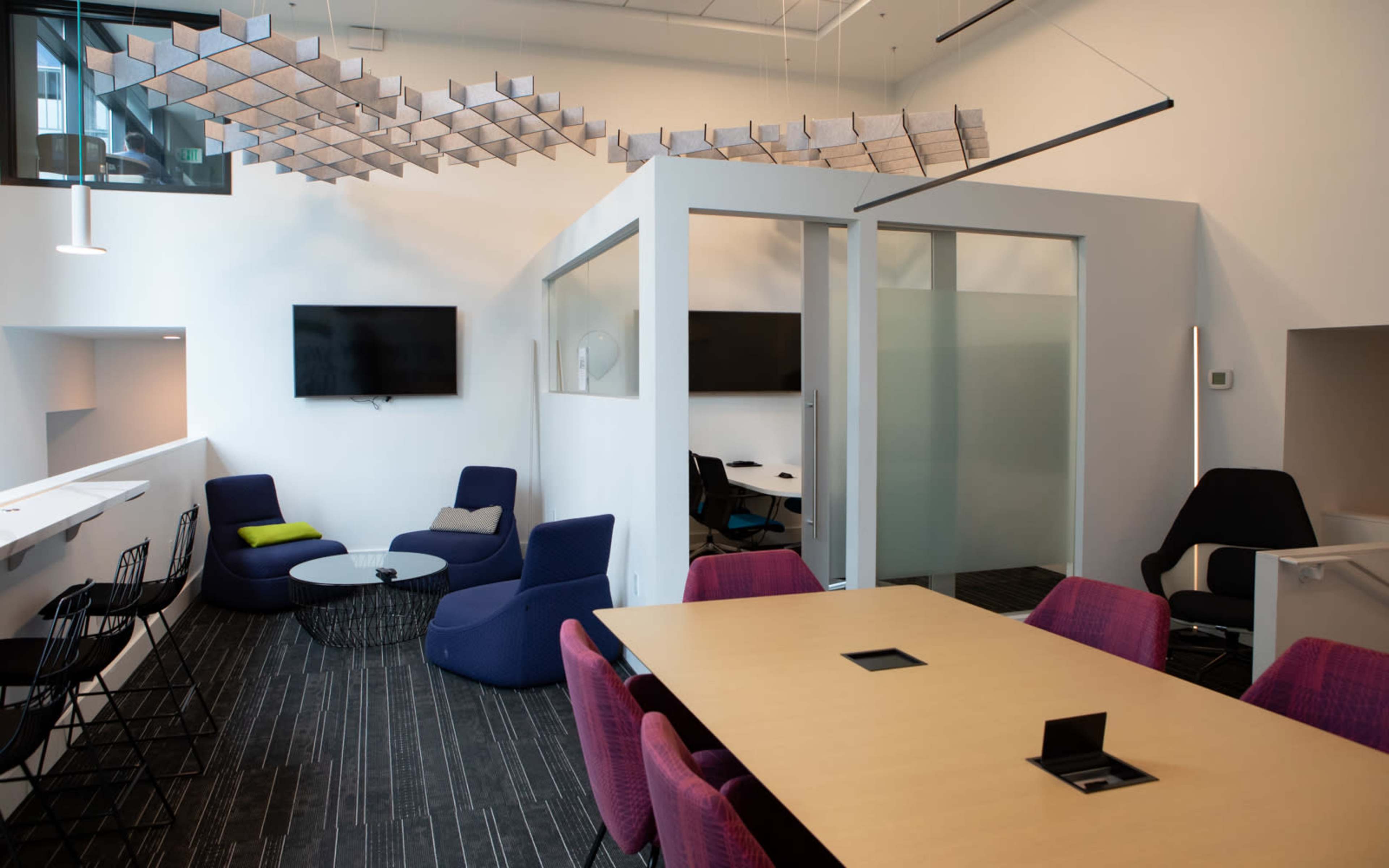 The image shows a modern office space featuring a mix of seating areas with colorful chairs and tables, alongside several enclosed meeting rooms.