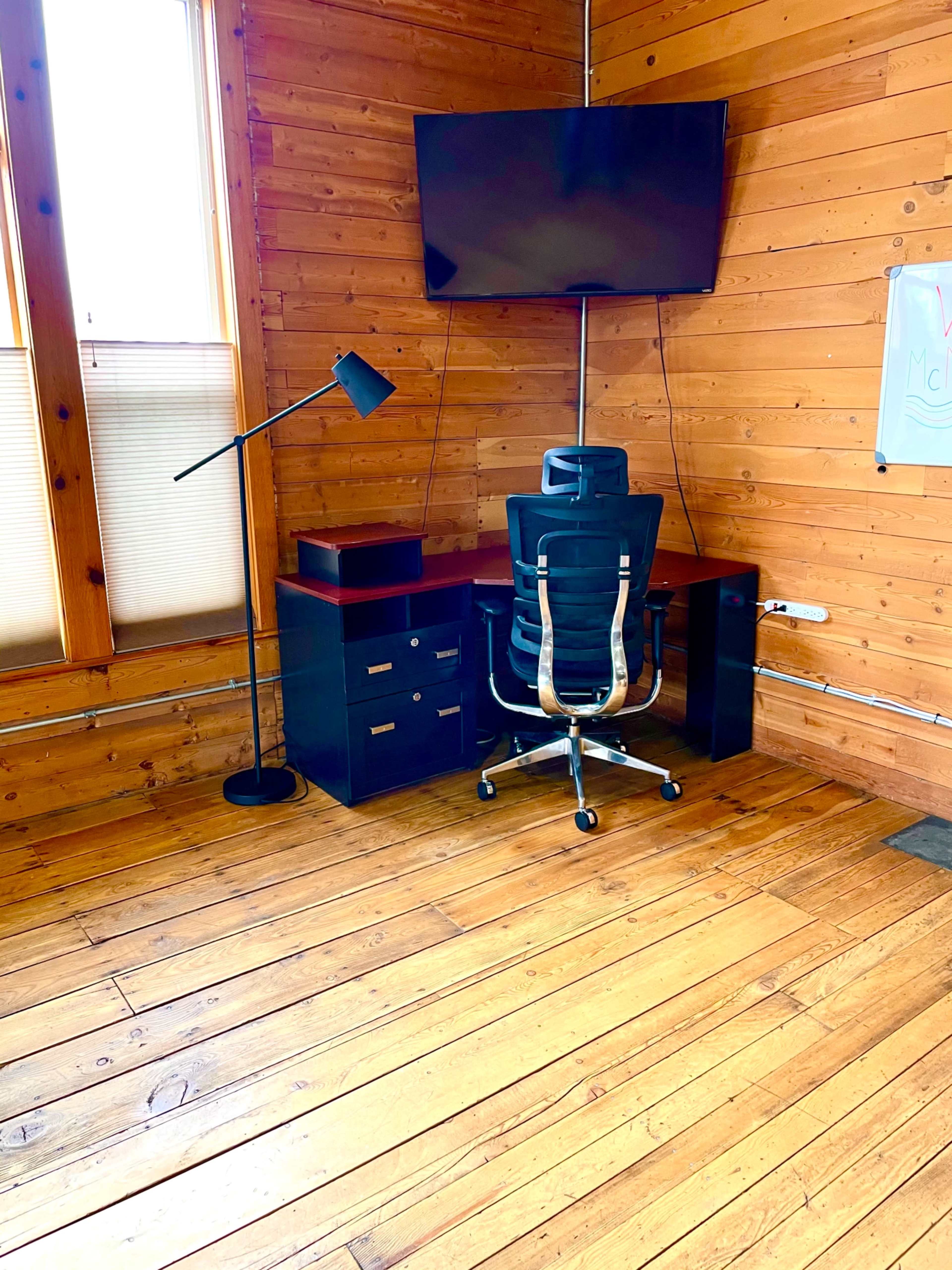 A wooden-floored room with a desk, an office chair, a television mounted on the wall, and a floor lamp.