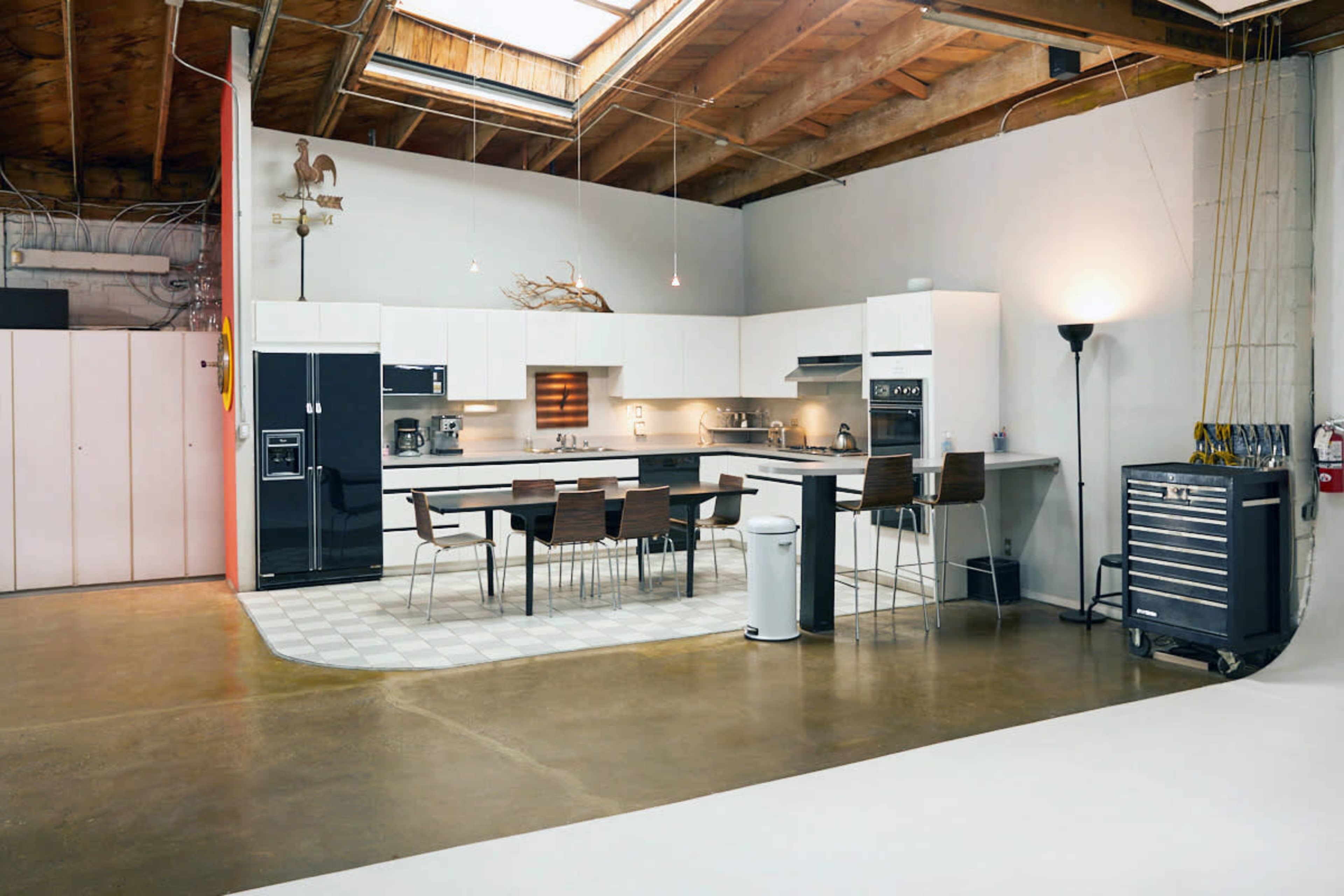 A modern kitchen with white cabinets, a large island with seating, and stainless steel appliances, set in a spacious, industrial-style room with exposed wooden beams.