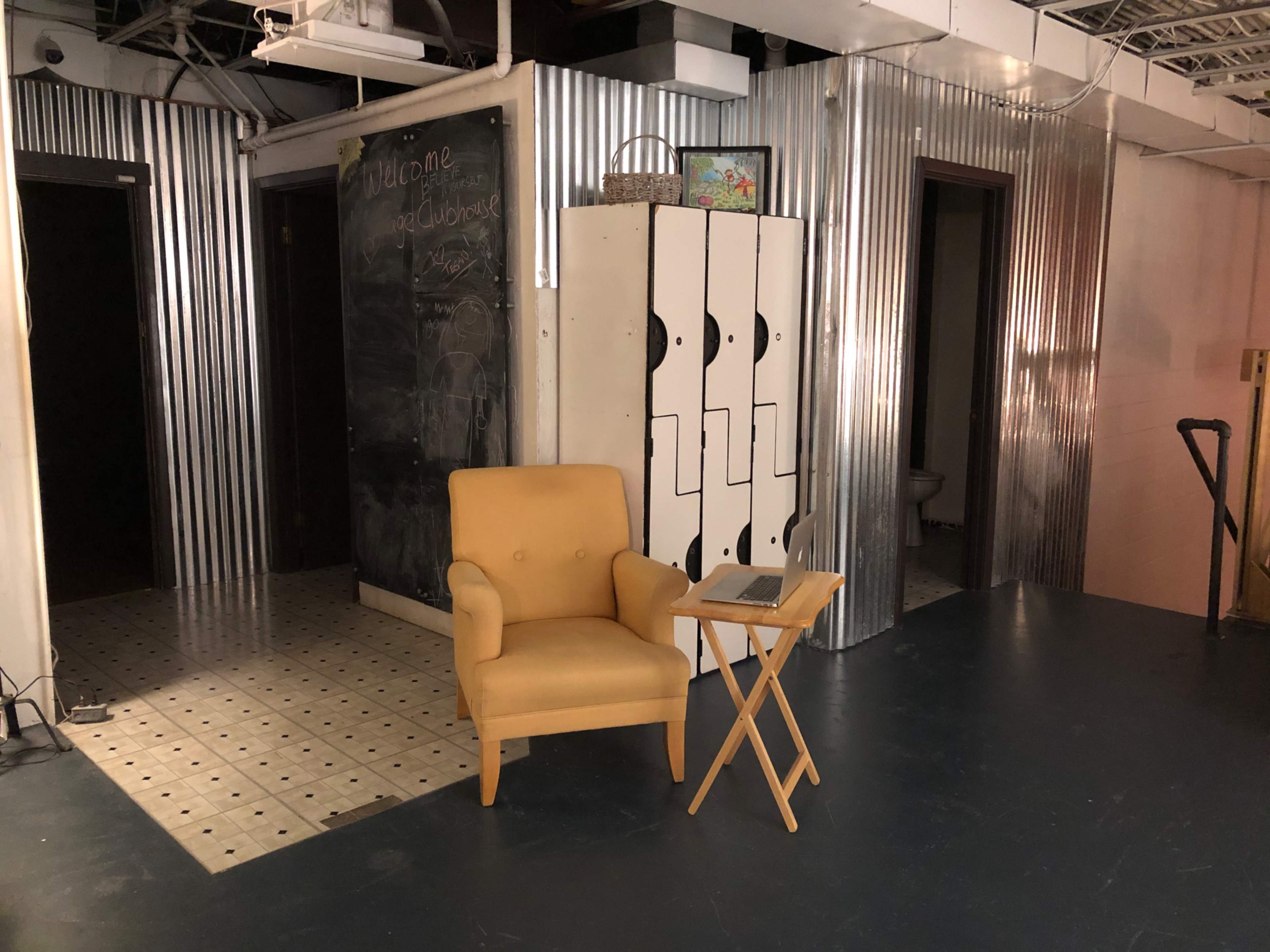 A yellow armchair next to a small folding table, set in a room with corrugated metal walls and two doorways.