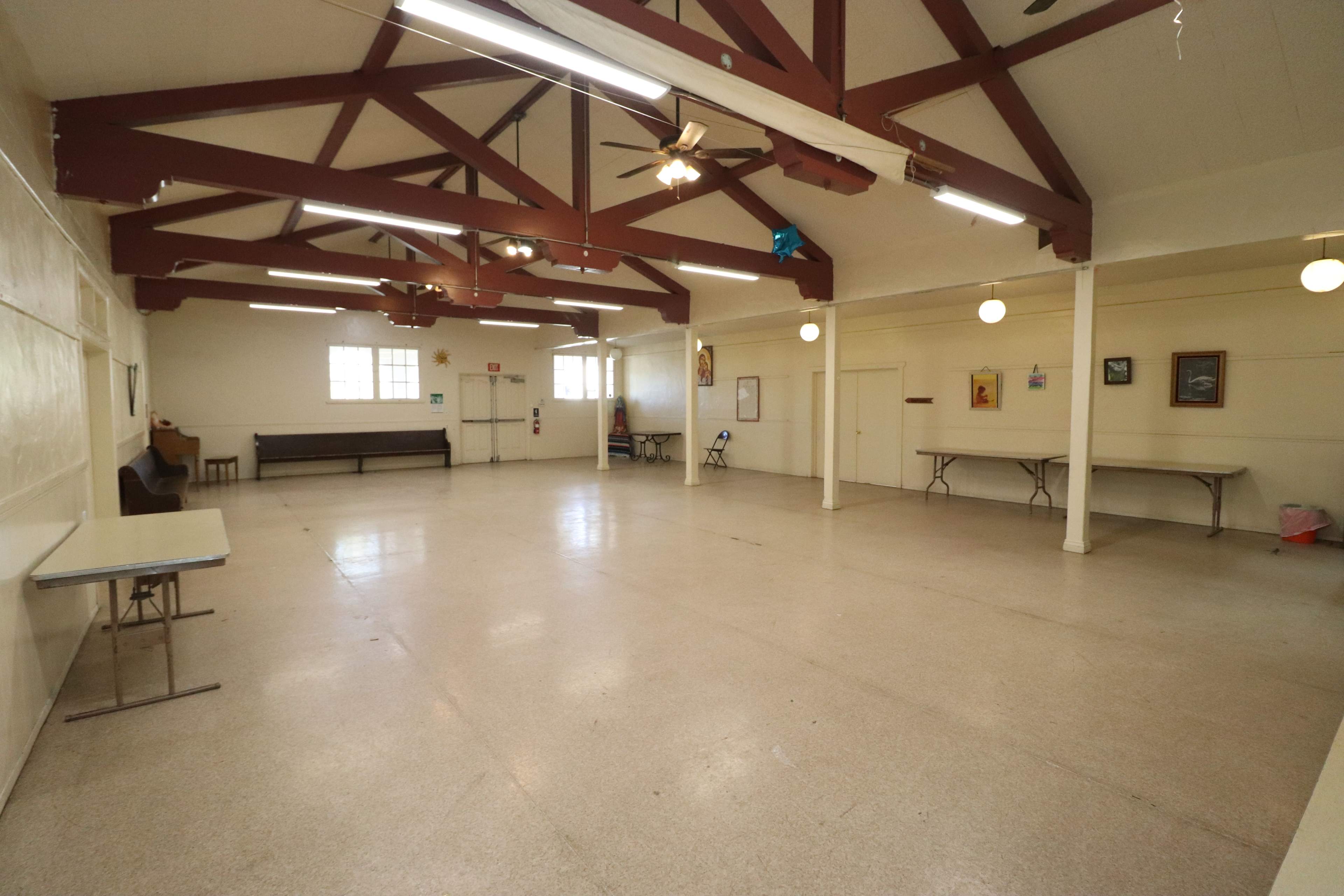 An empty hall with a high ceiling, wooden beams, and a few tables and chairs scattered around.