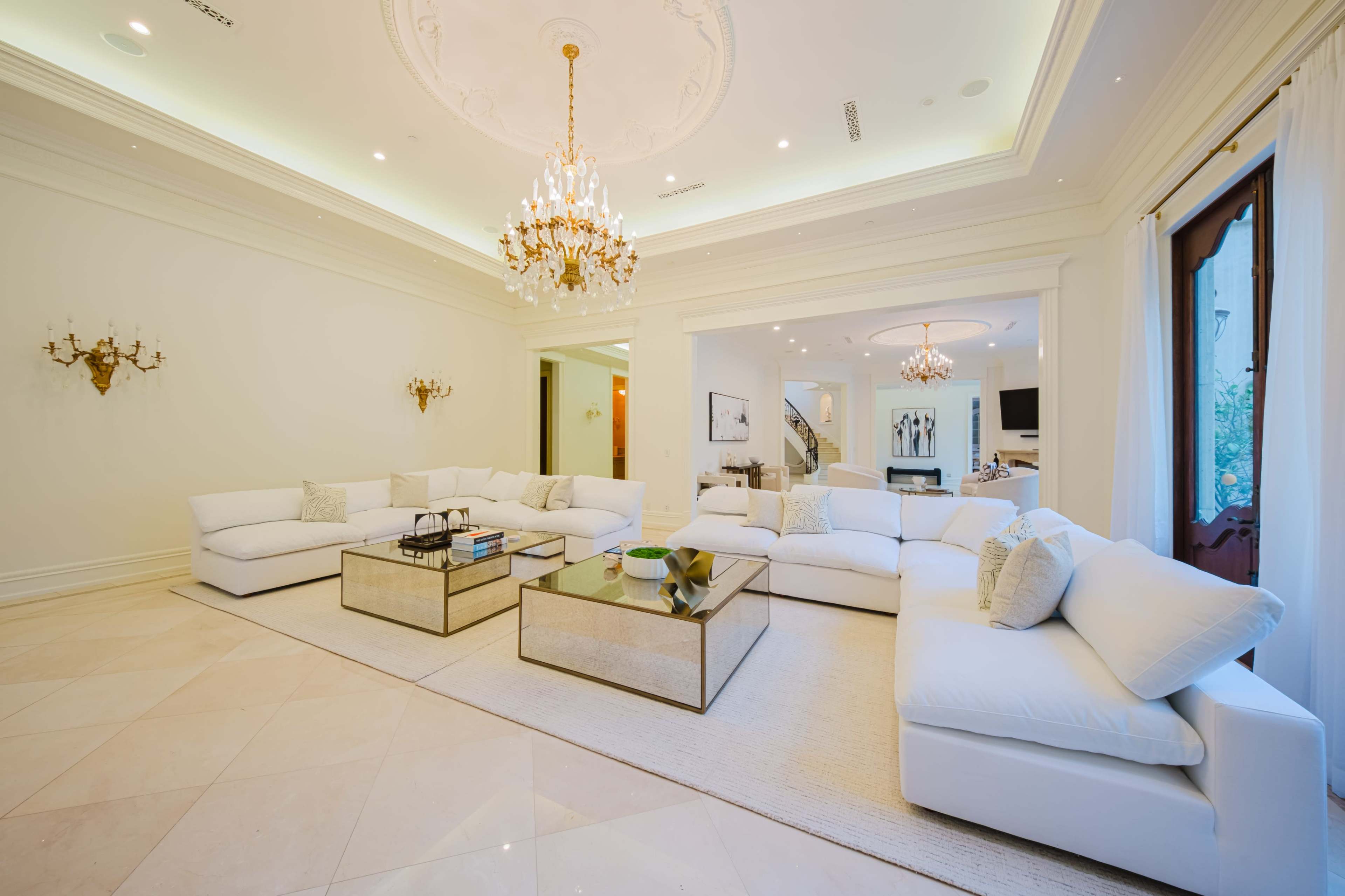 The image shows a spacious living room with white furnishings, a large area rug, and two elegant chandeliers hanging from the ceiling.