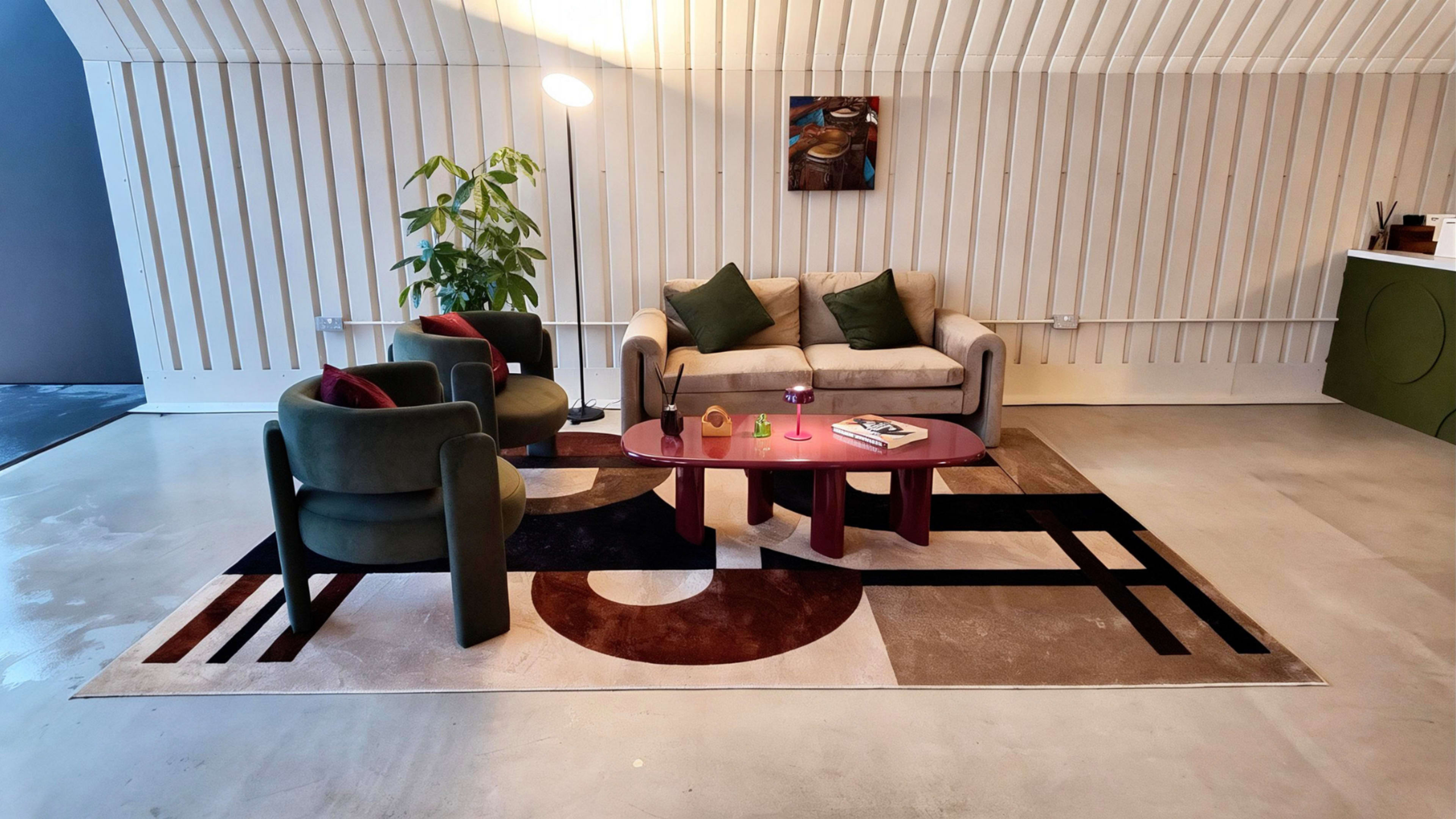 A modern living room features a beige sofa, two green armchairs, a round coffee table, and a patterned area rug against a backdrop of wooden slatted walls.