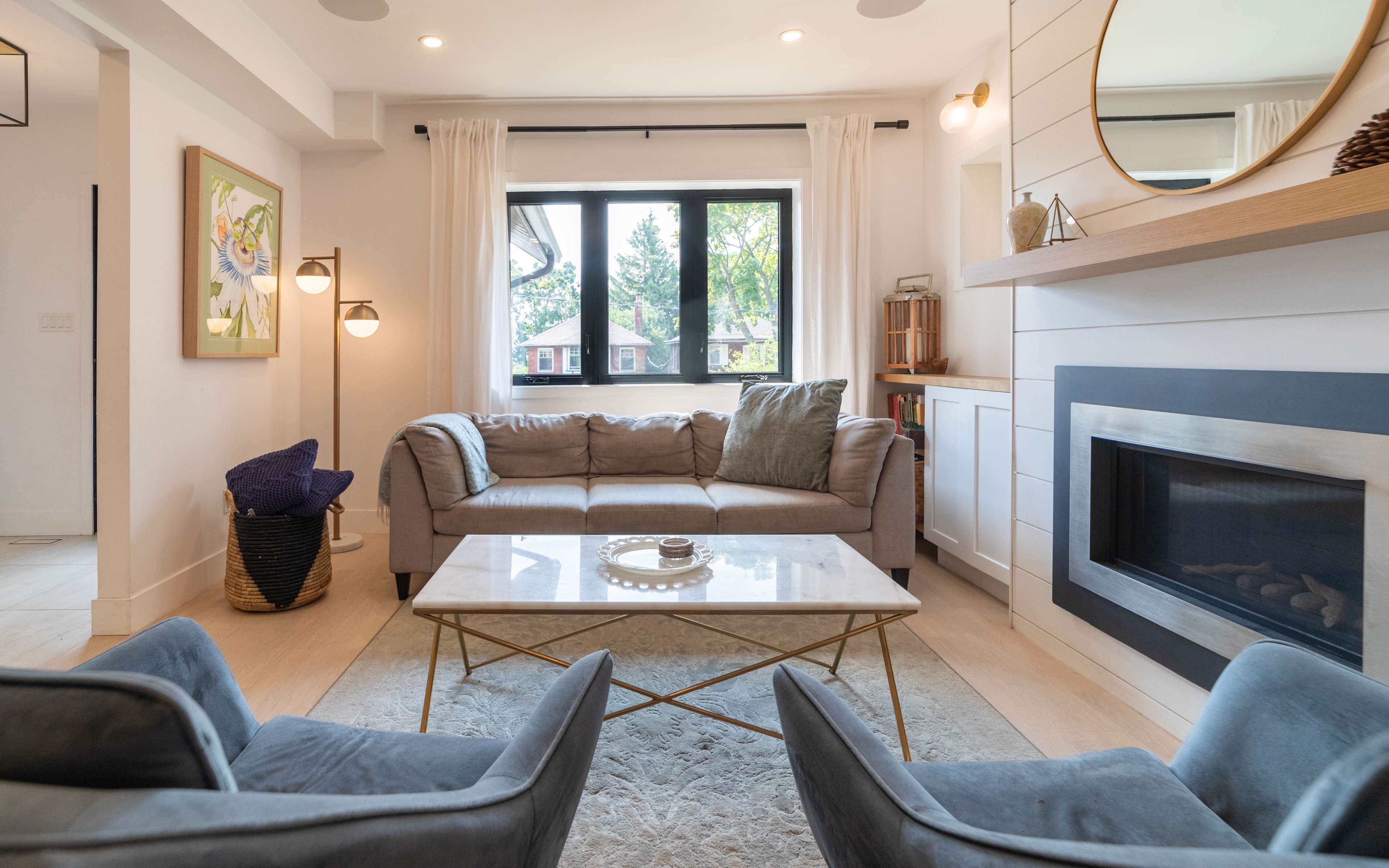 A contemporary living room features a gray sofa and two blue chairs facing a glass coffee table, with large windows allowing natural light to enter the space.
