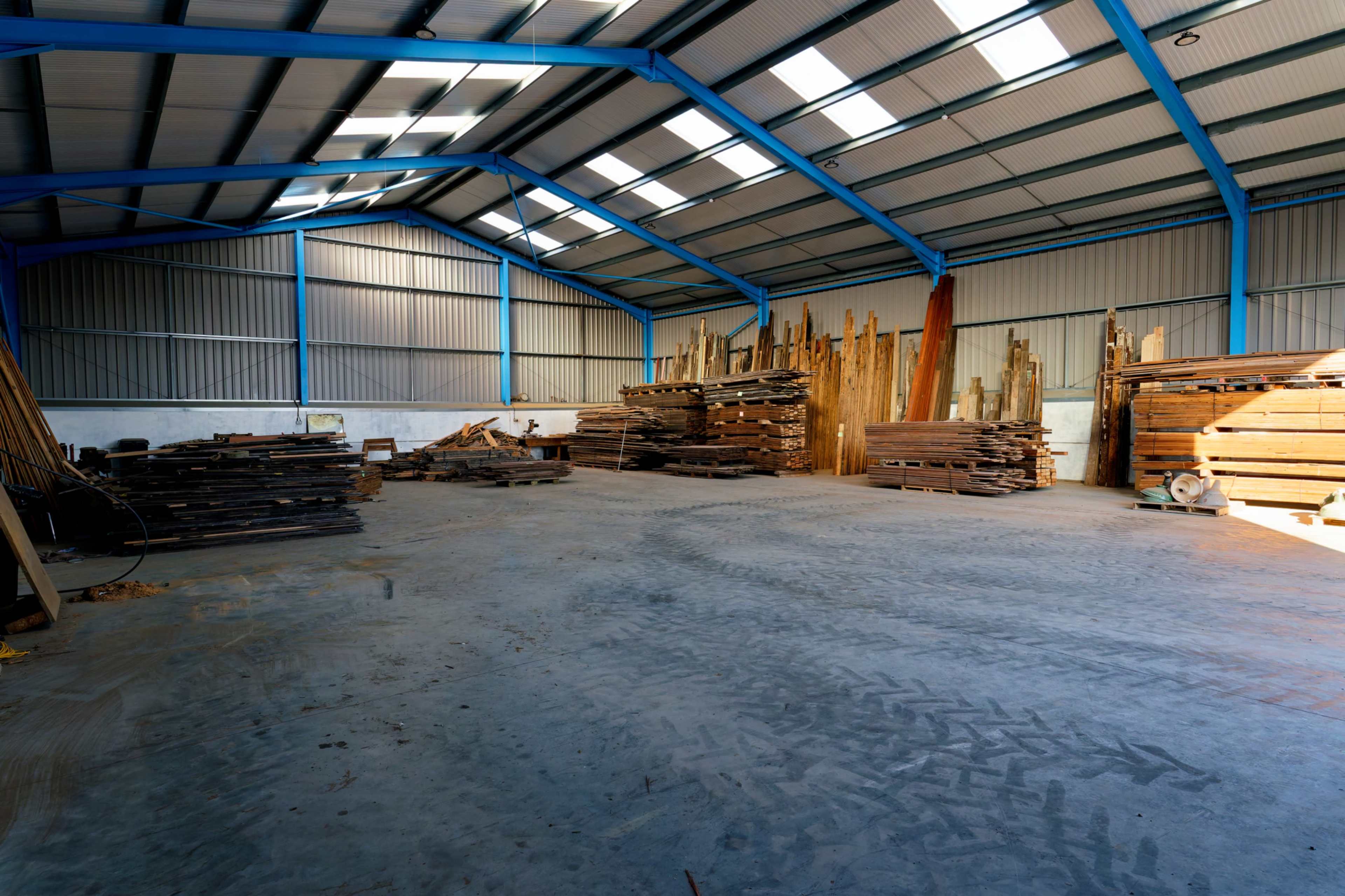 The image shows an industrial warehouse with stacks of lumber and wooden pallets arranged on the concrete floor under a metal roof.