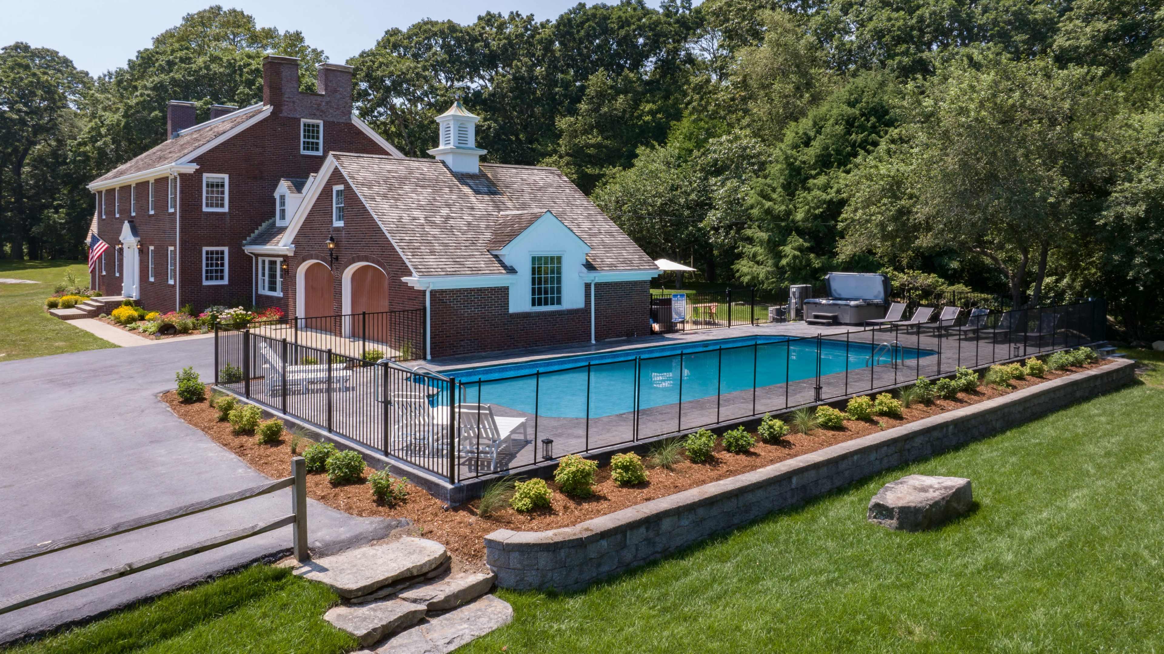 The image shows a large brick house with a gabled roof next to a rectangular swimming pool surrounded by a black metal fence and landscaped with grass and flower beds.