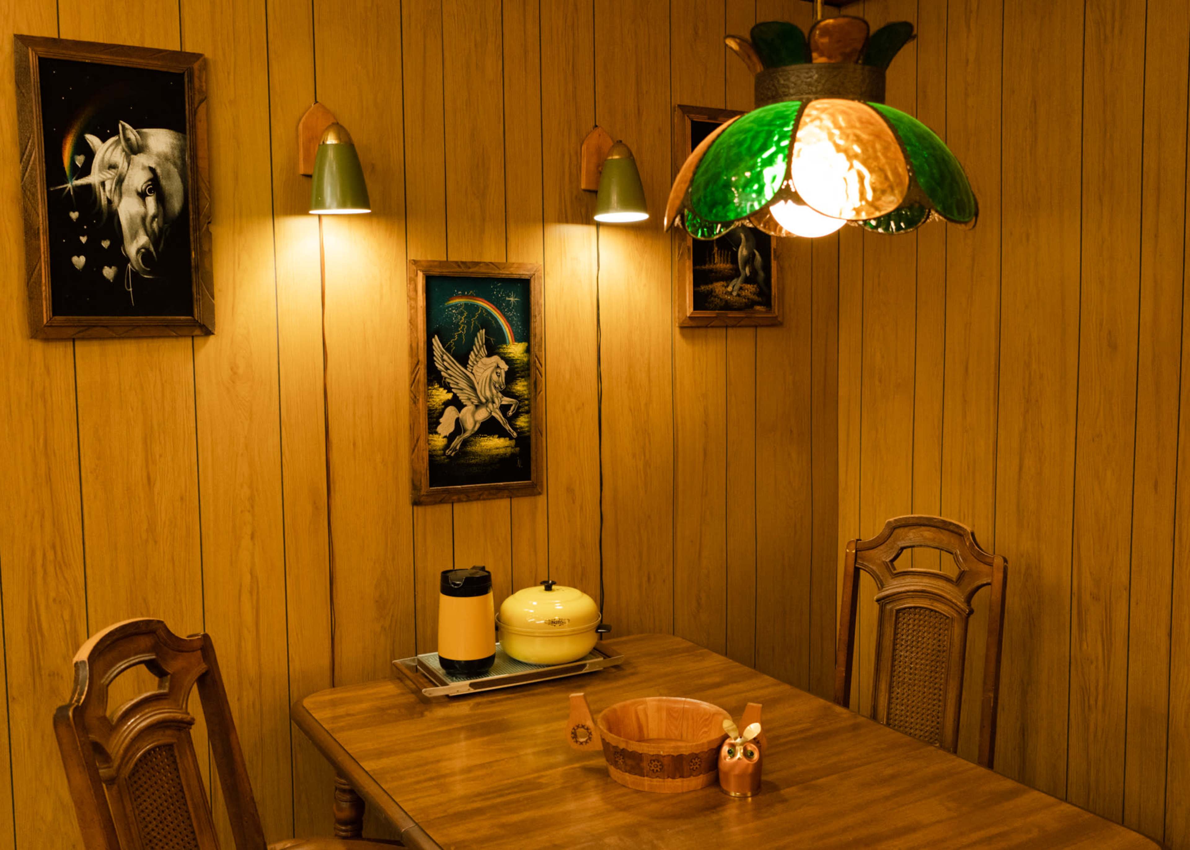 The image shows a cozy dining area with wooden paneling, a table set for meals, and a green stained glass lamp hanging above.