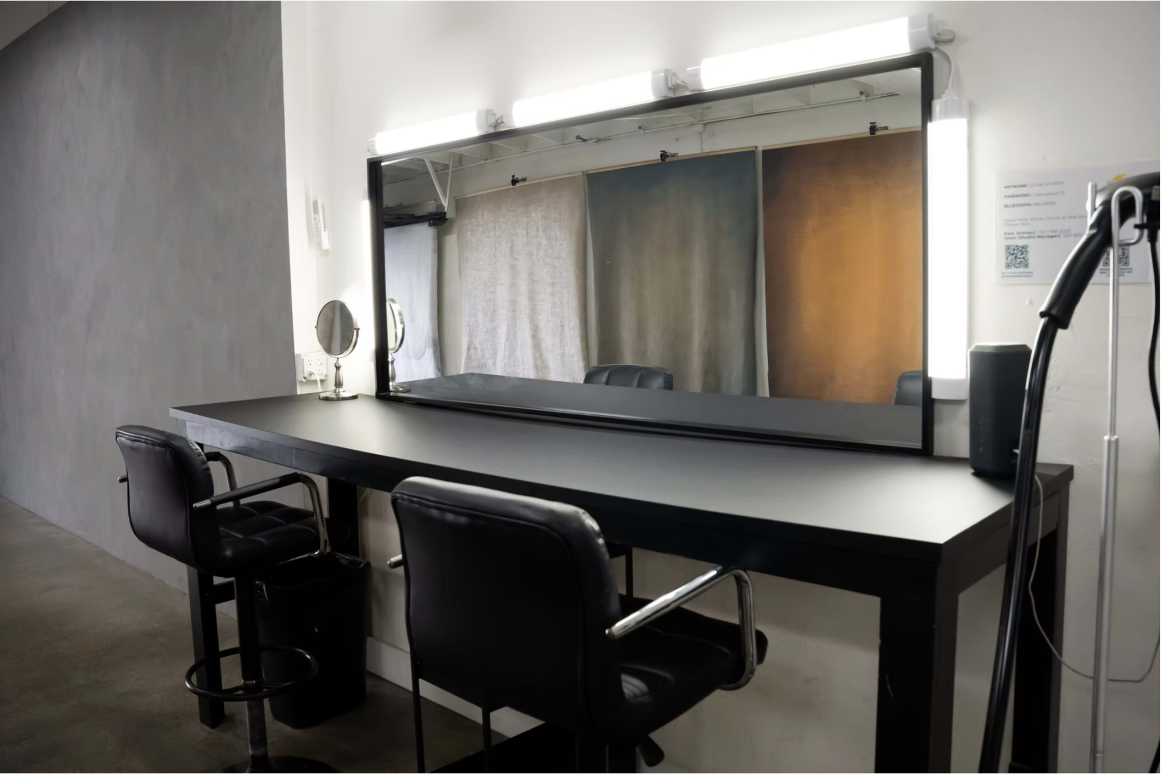 The image shows a minimalist salon space with a black countertop, two black chairs, and a large mirror illuminated by overhead lights.