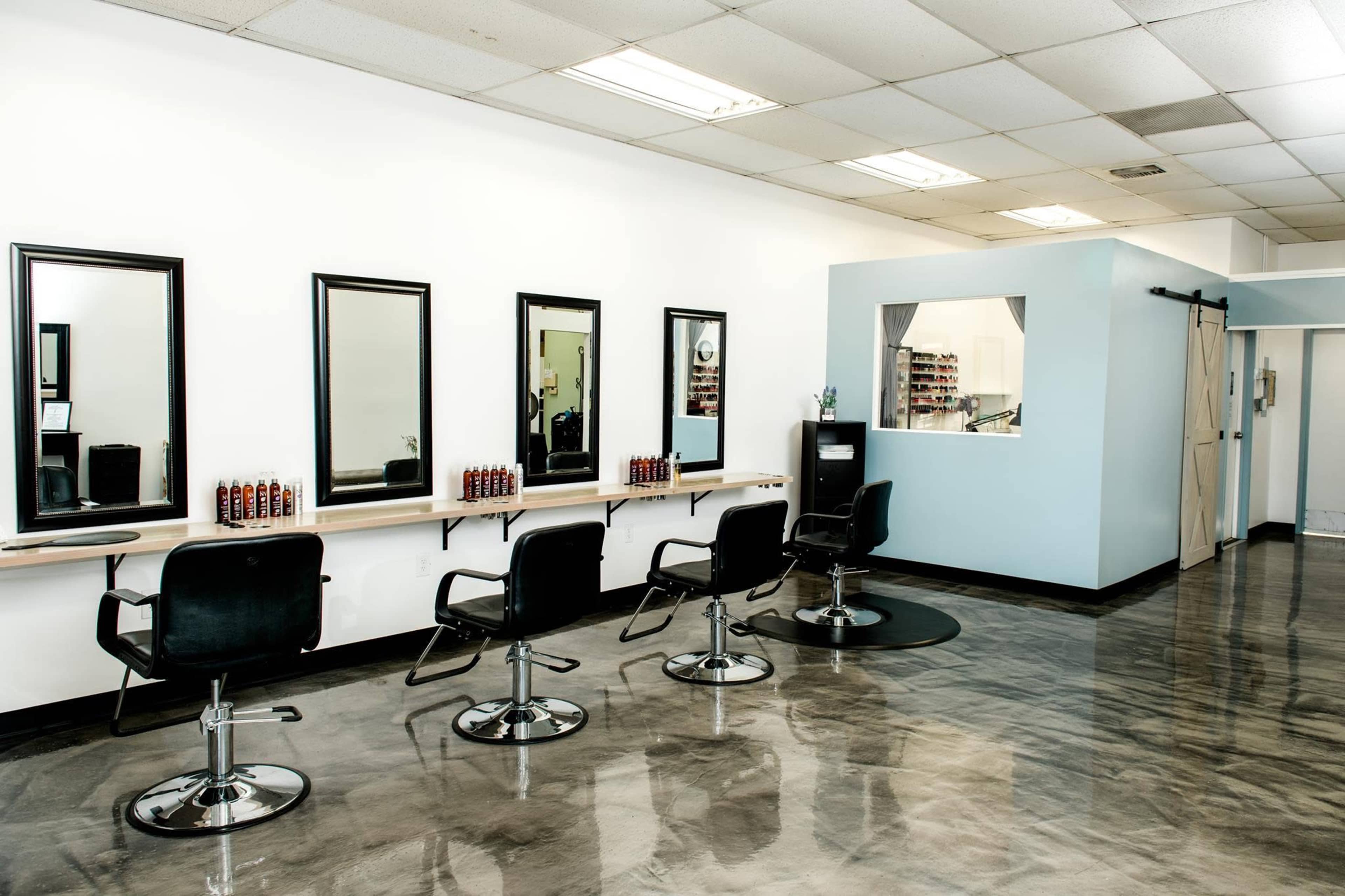 The image shows a modern hair salon with four black styling chairs in front of mirrors, a waiting area, and a reception desk against a light blue wall.