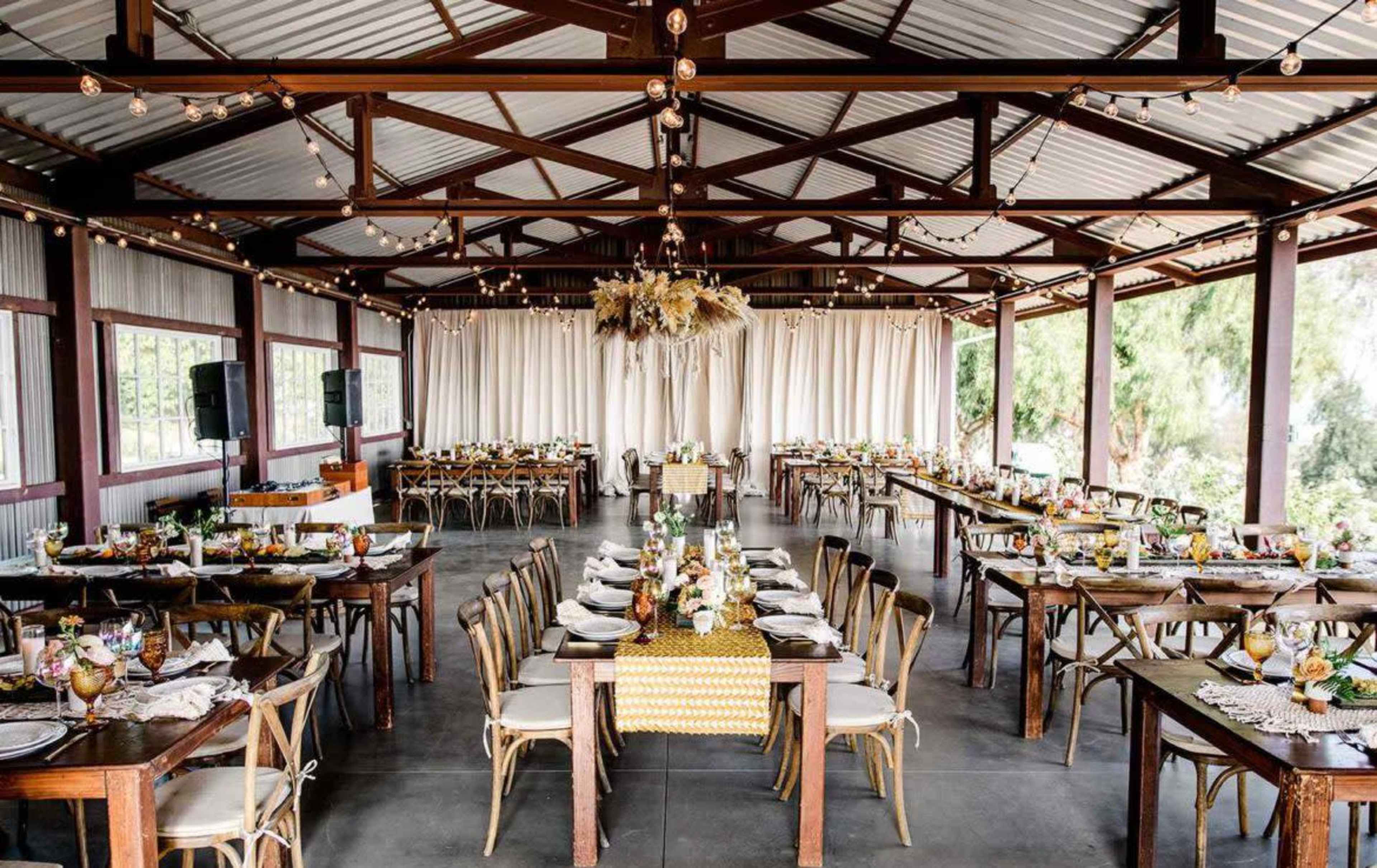 A spacious event venue features rows of wooden tables set with tableware, surrounded by a fabric-draped backdrop and hanging string lights.