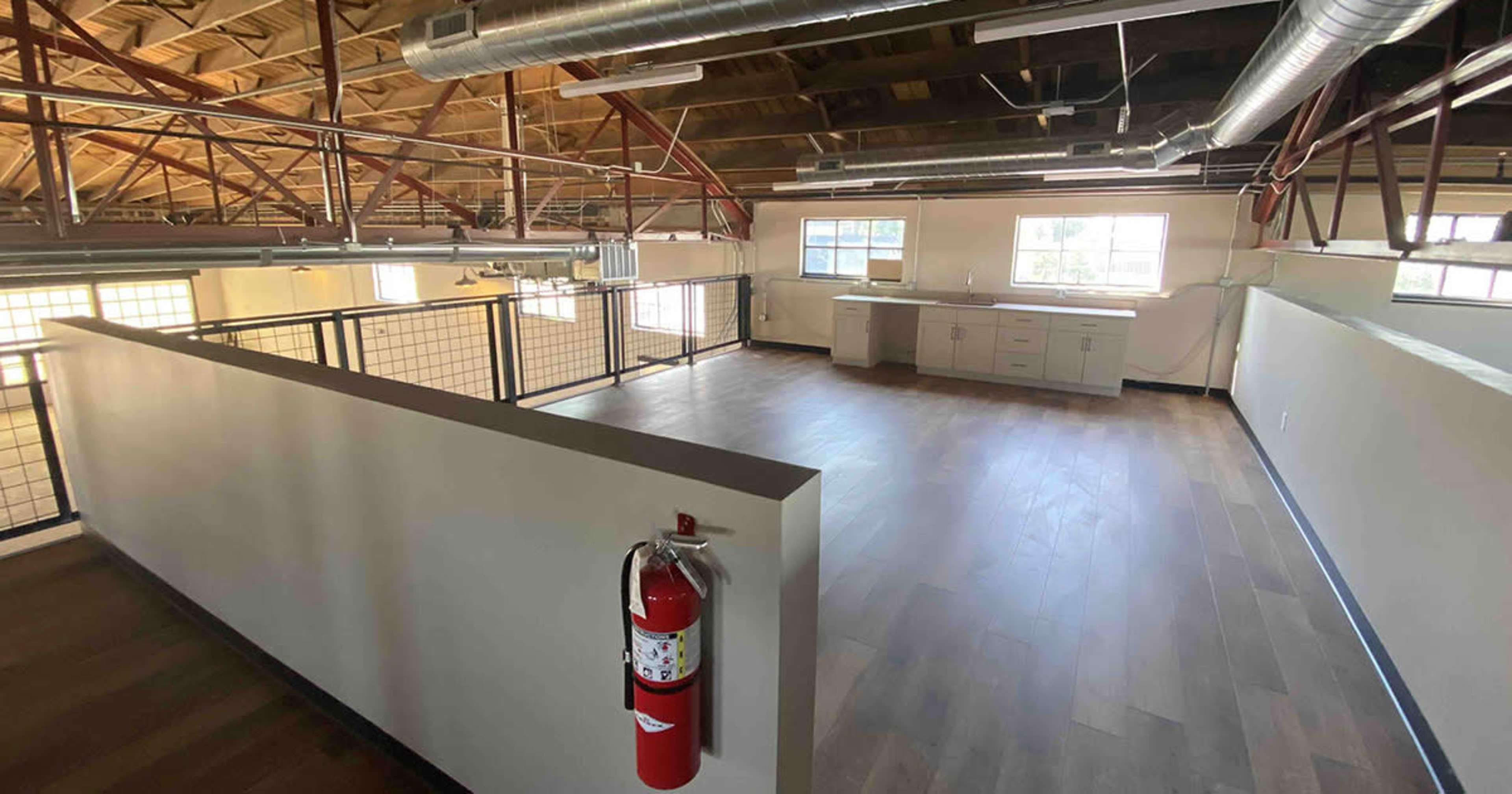 The image shows an open, brightly lit loft space with wooden floors, large windows, and a small kitchenette area.