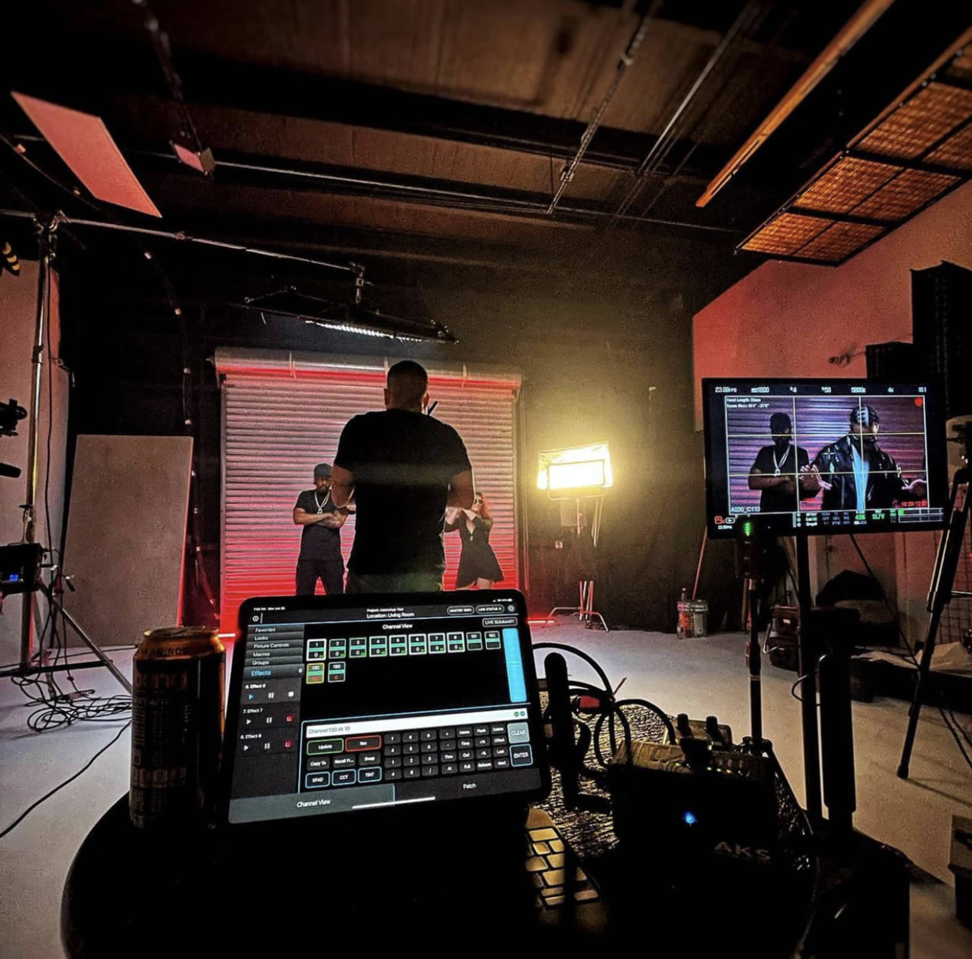 A film or photography studio with a person operating equipment at a control panel while two individuals perform in front of a backdrop.
