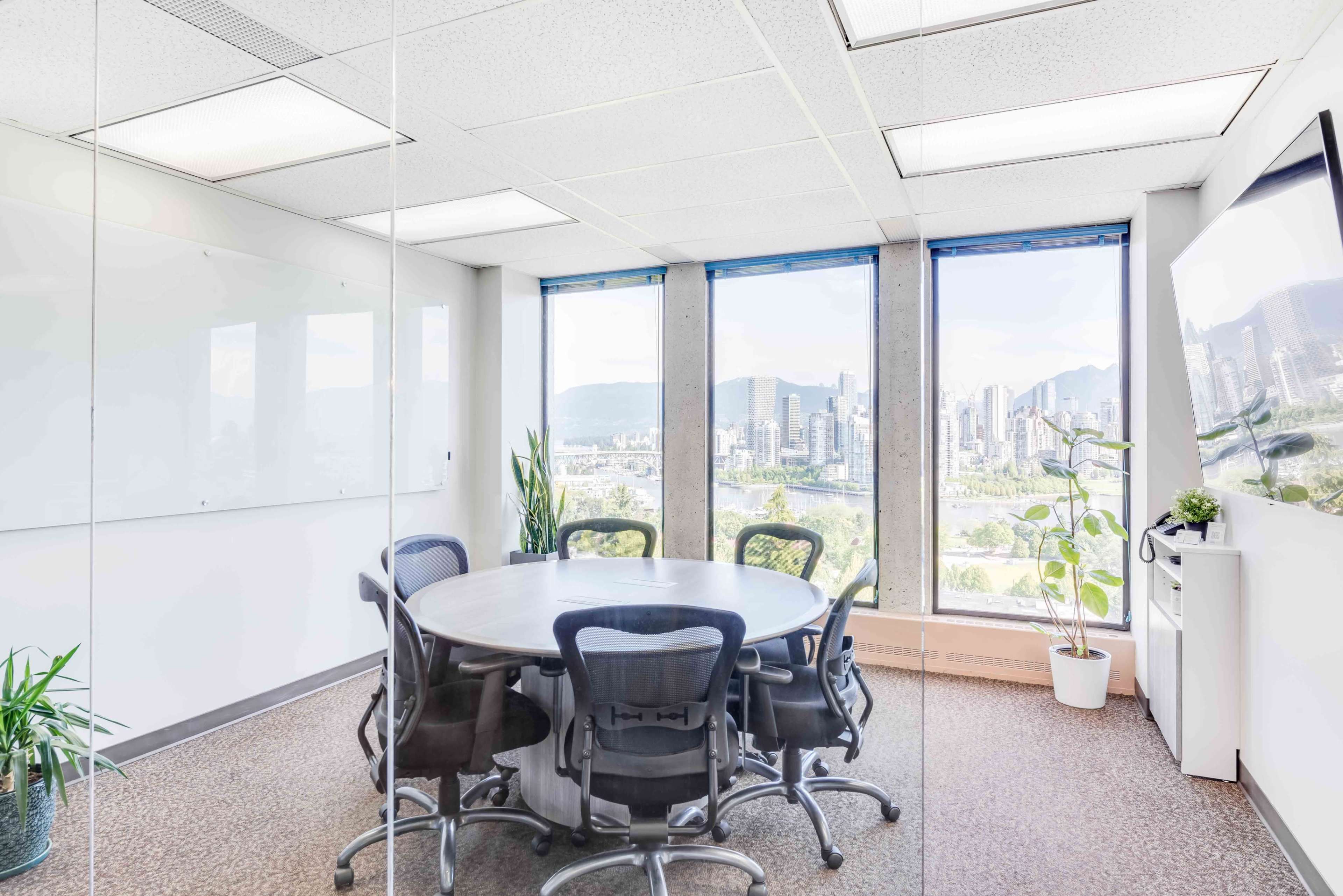A conference room with a round table and chairs overlooks a cityscape through large windows.