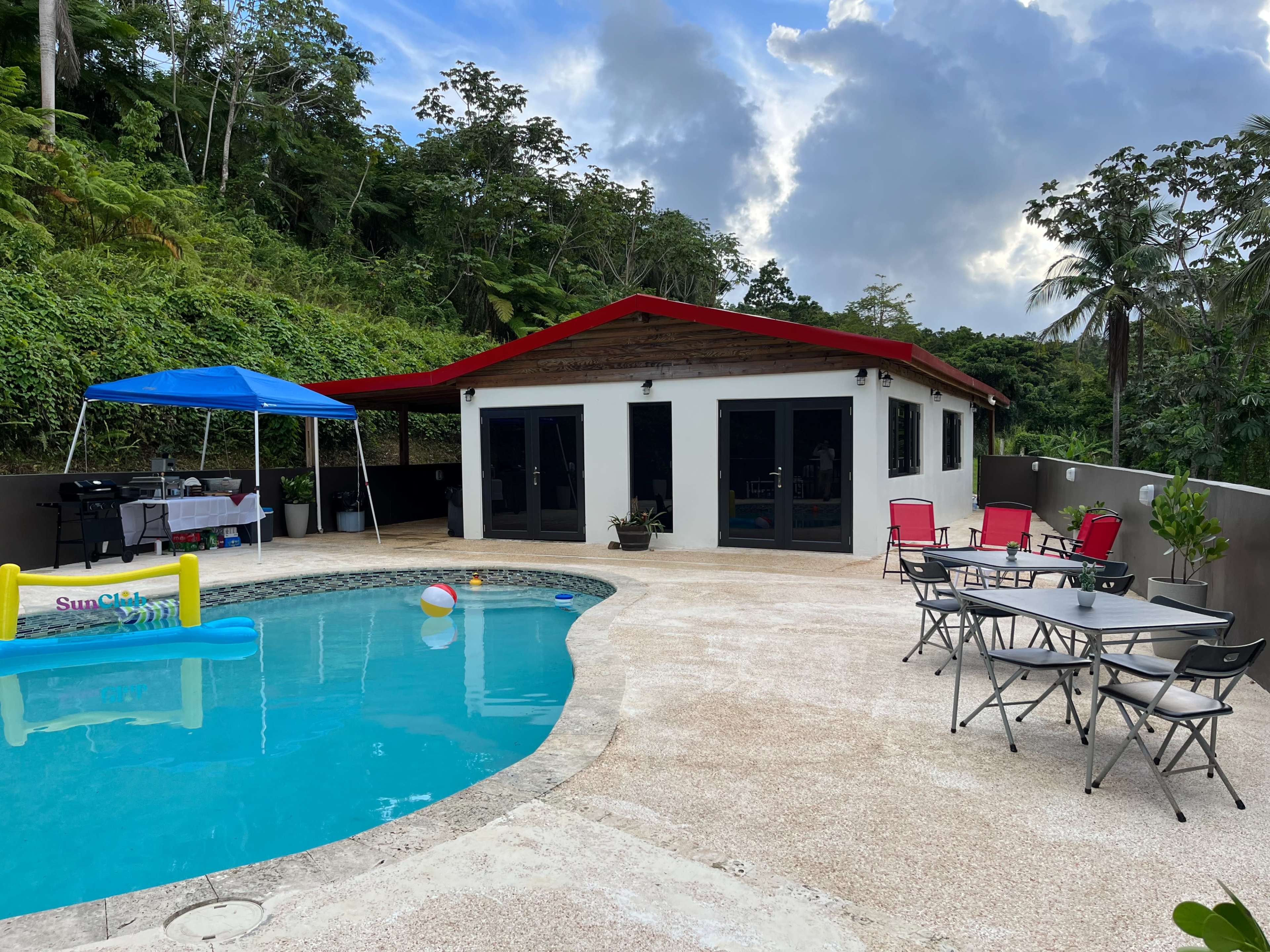 A modern house with a red roof is situated next to a swimming pool surrounded by lounge chairs and a shaded seating area.