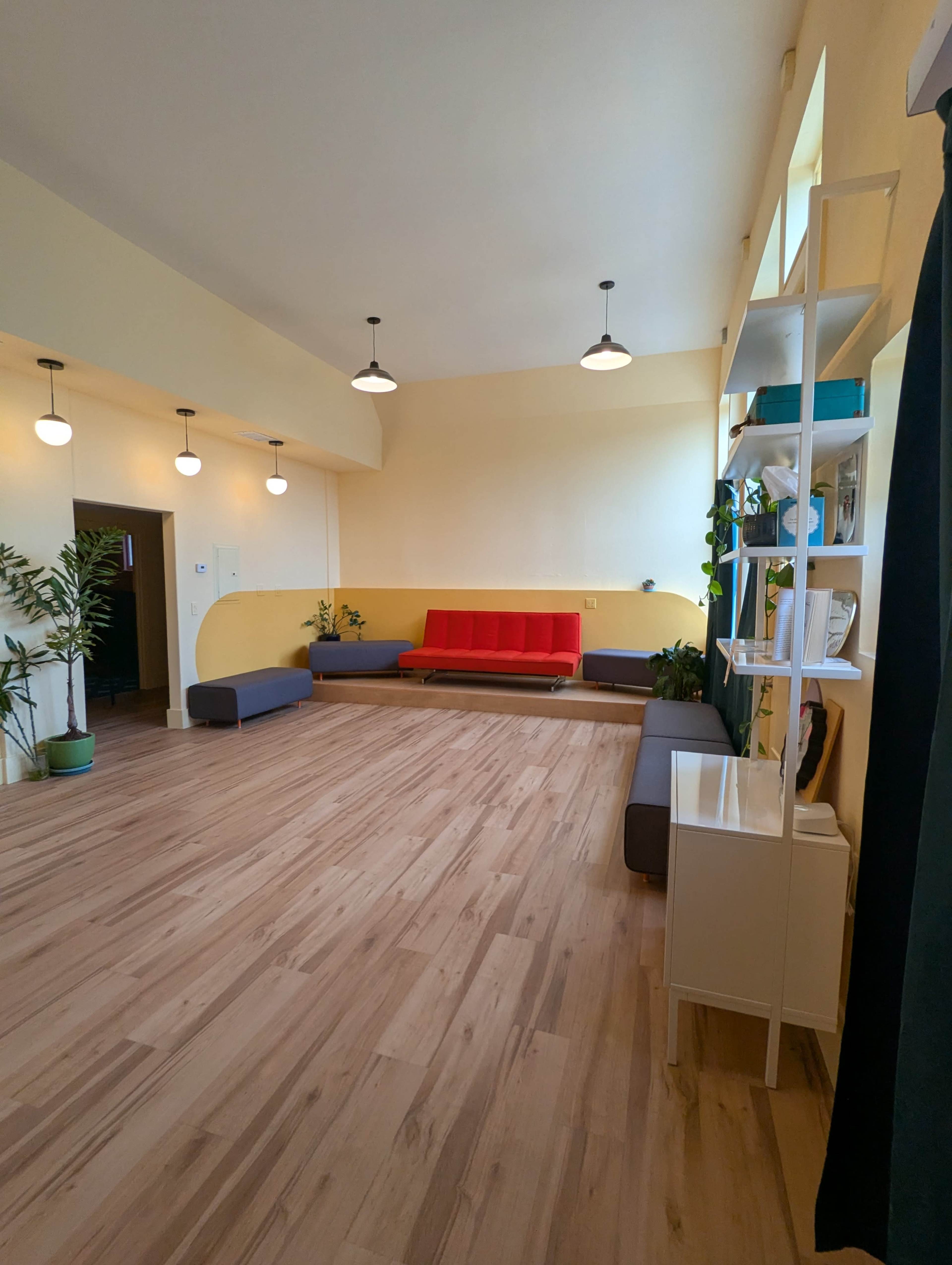 The image shows a spacious room with yellow walls, wooden flooring, a red sofa, gray seating, and plants arranged throughout.