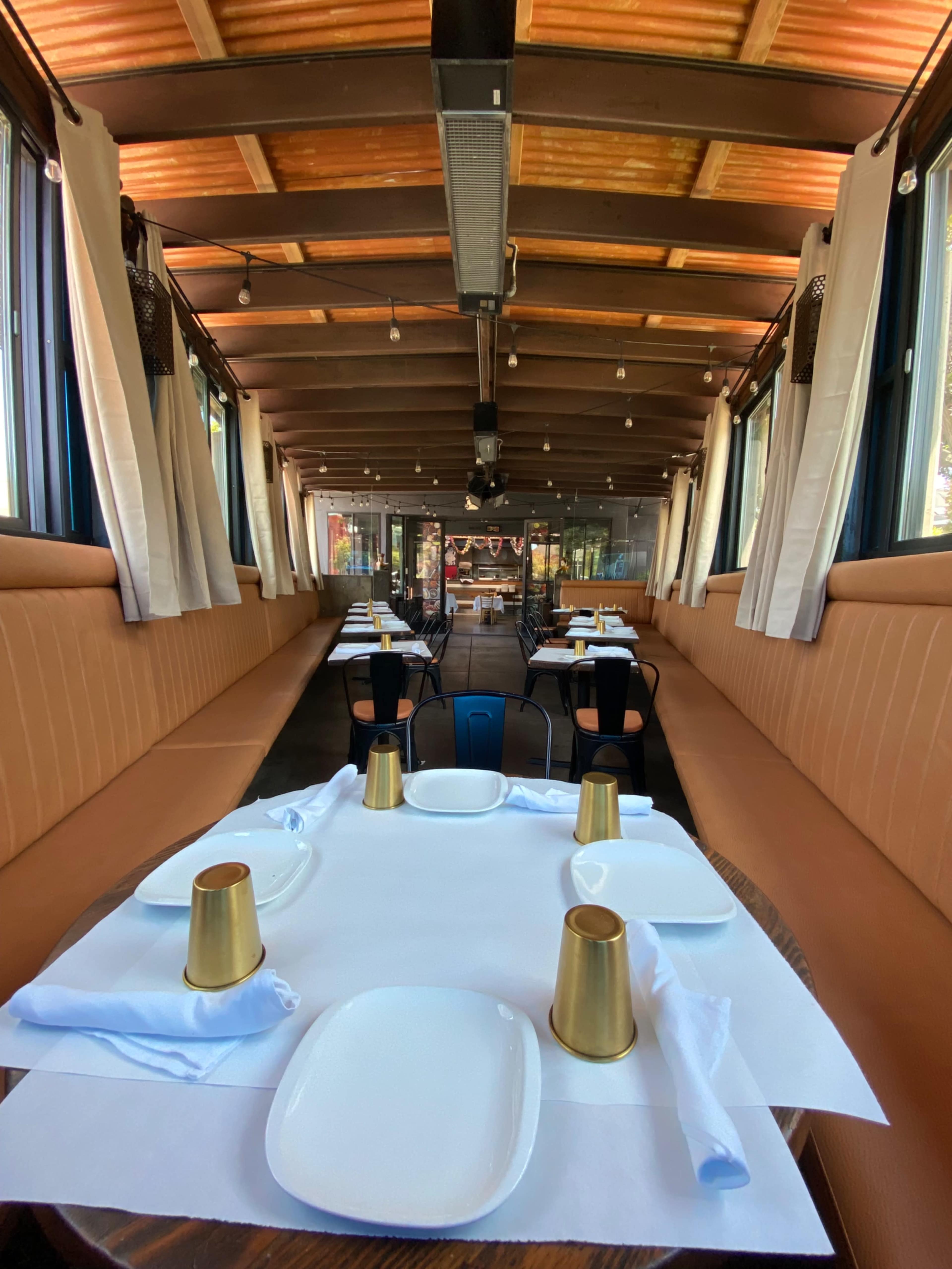 A restaurant interior featuring tables set with white tablecloths and gold cups under a wooden ceiling with large windows.