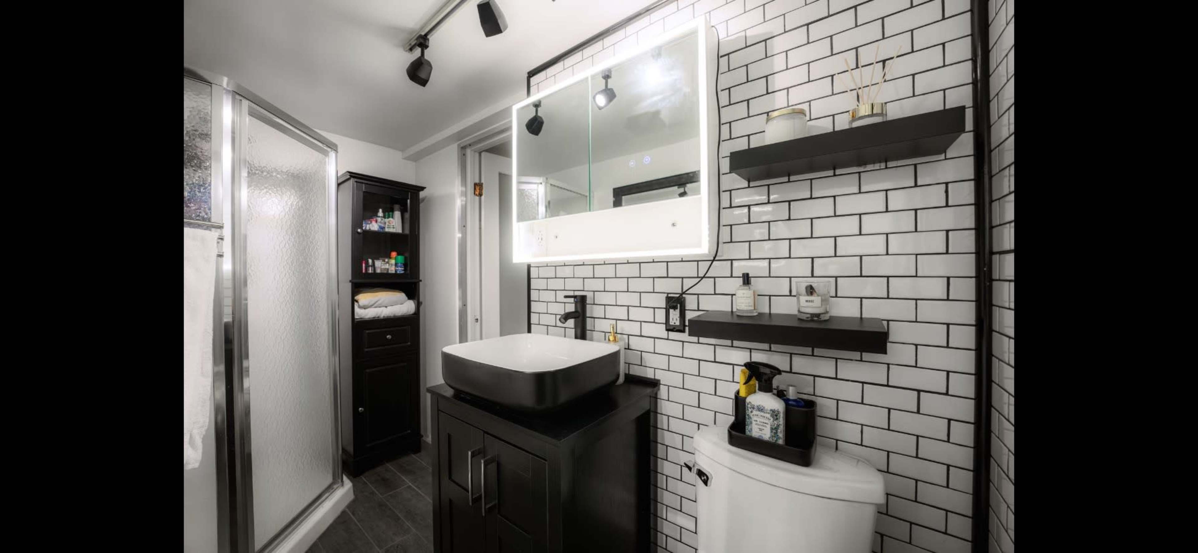 A modern bathroom featuring a black vanity with a rectangular sink, a mirrored cabinet above, and tiled walls comprising black and white subway tiles.