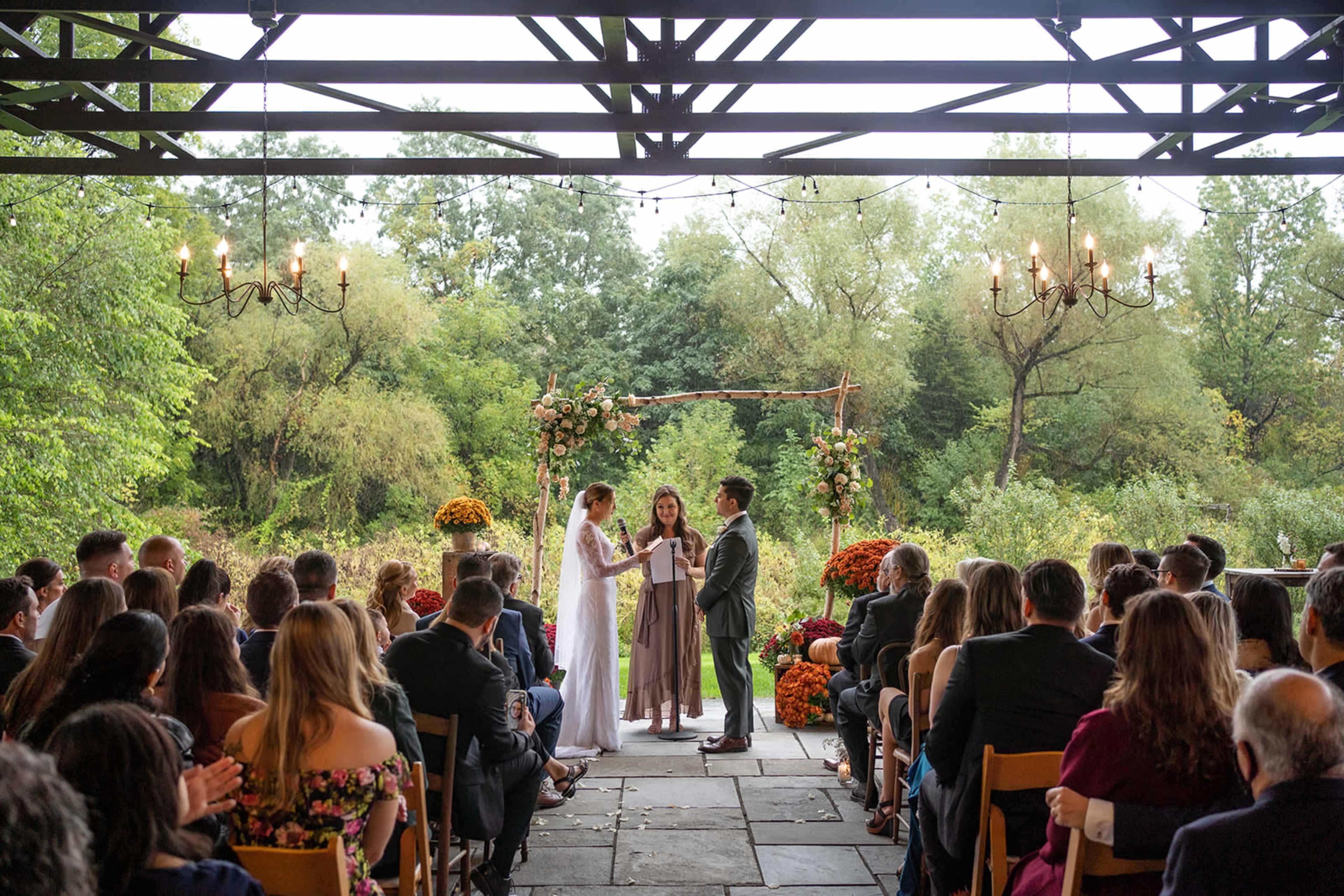 A wedding ceremony takes place outdoors under a wooden canopy, surrounded by seated guests and decorated with flowers and autumn foliage.