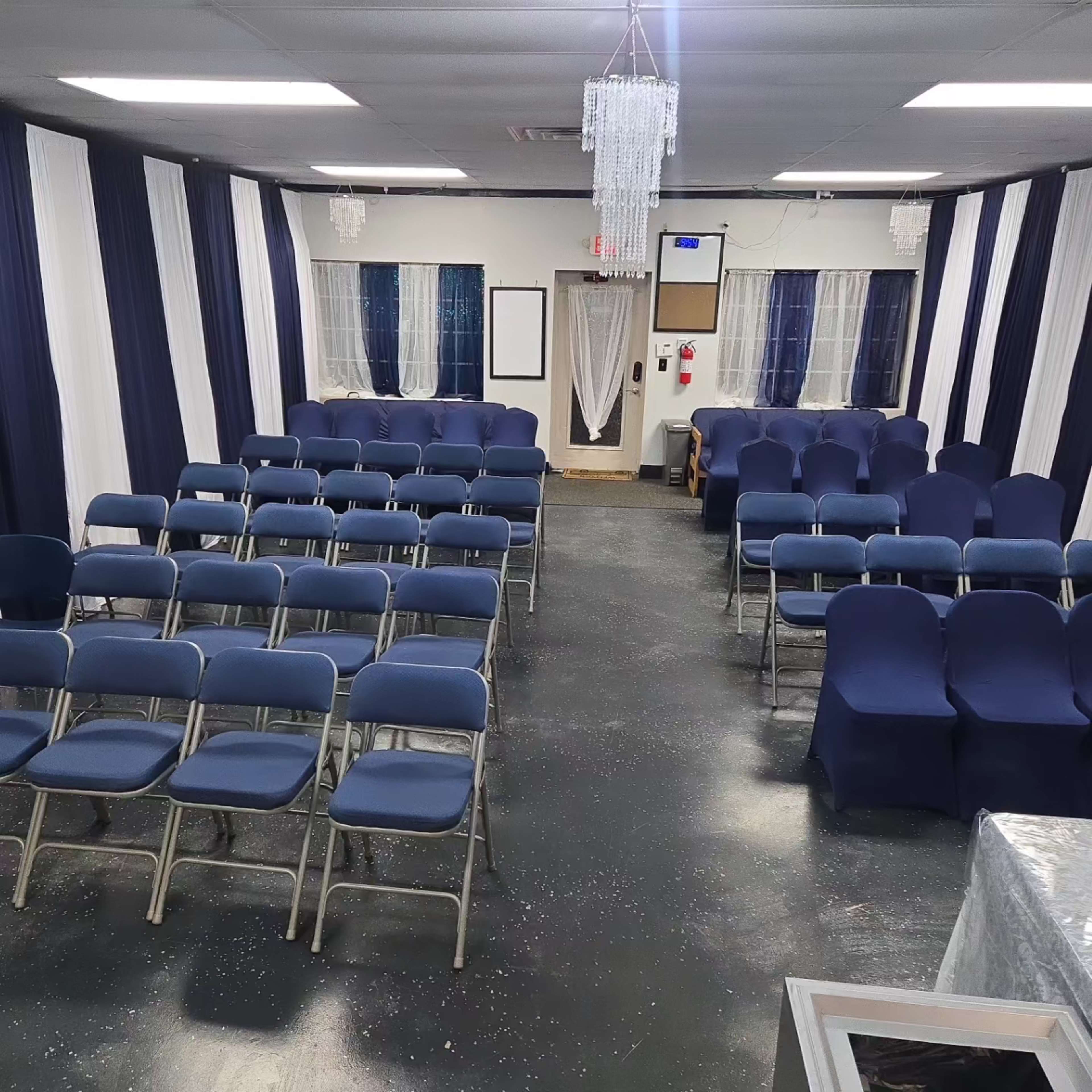 The image shows a room set with rows of blue chairs and neatly draped fabric on the walls, featuring a small stage area and decorative lighting.